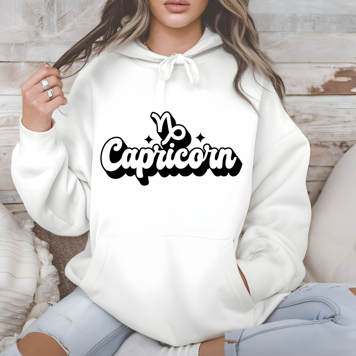 A woman wearing a white pullover hoodie with a black 'Yo Capricorn' graphic on the chest, sitting against a rustic wood panel background.
