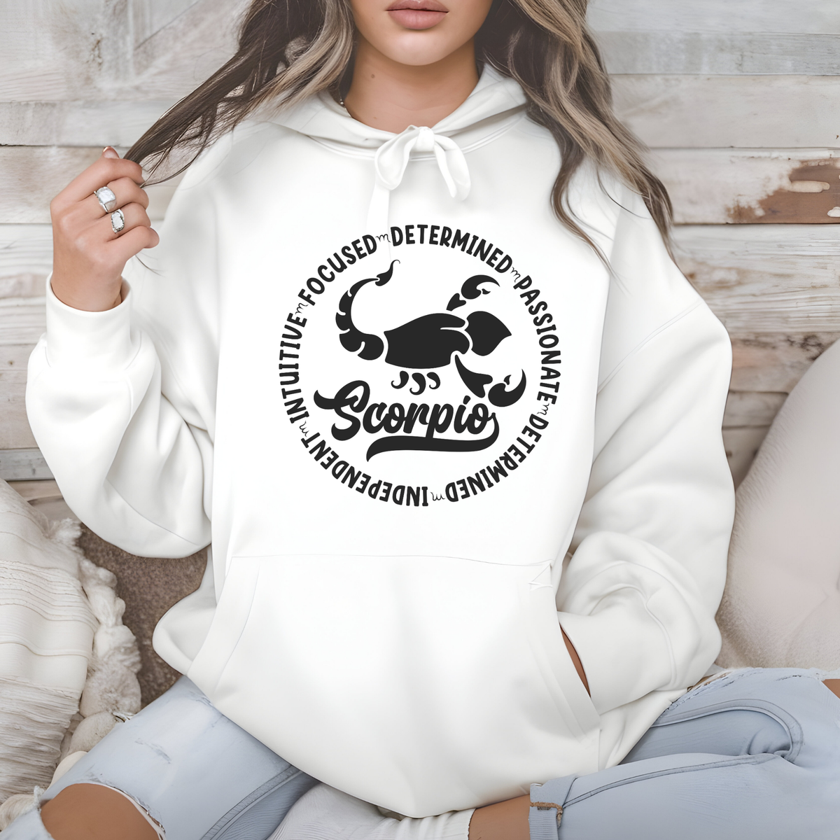Woman wearing a white crewneck sweatshirt featuring a white circular Scorpio zodiac graphic with a scorpion illustration, the word "Scorpio," and trait keywords like intuitive, focused, determined, passionate, and independent.