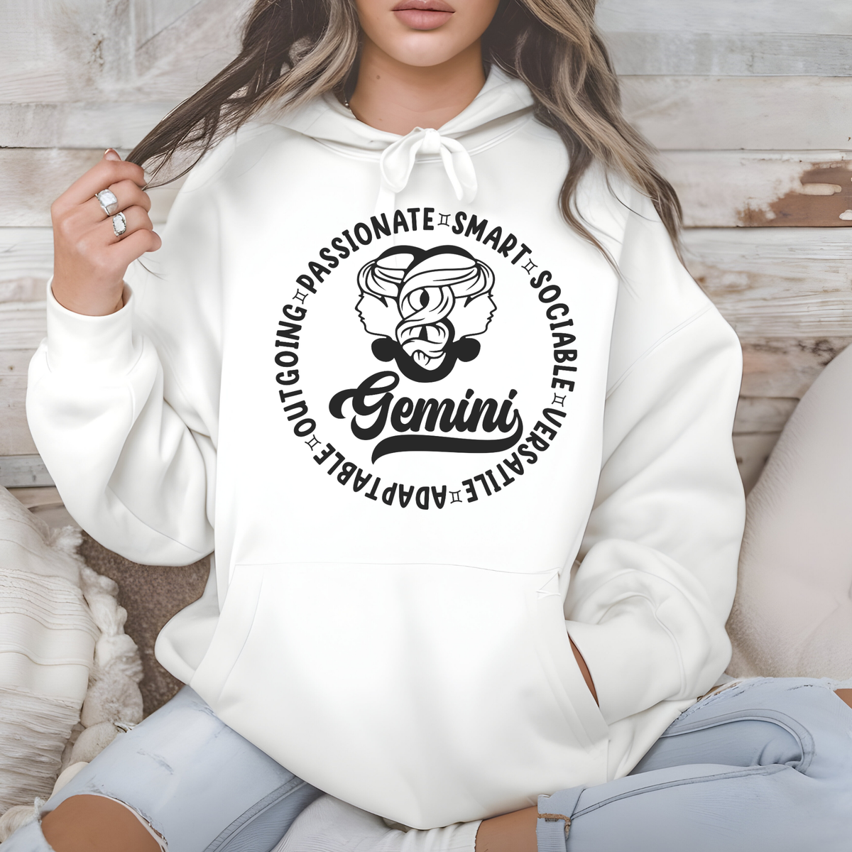 A woman wearing a white pullover hoodie with a black Gemini zodiac graphic on the chest, sitting in a cozy indoor setting with a rustic wood wall background.