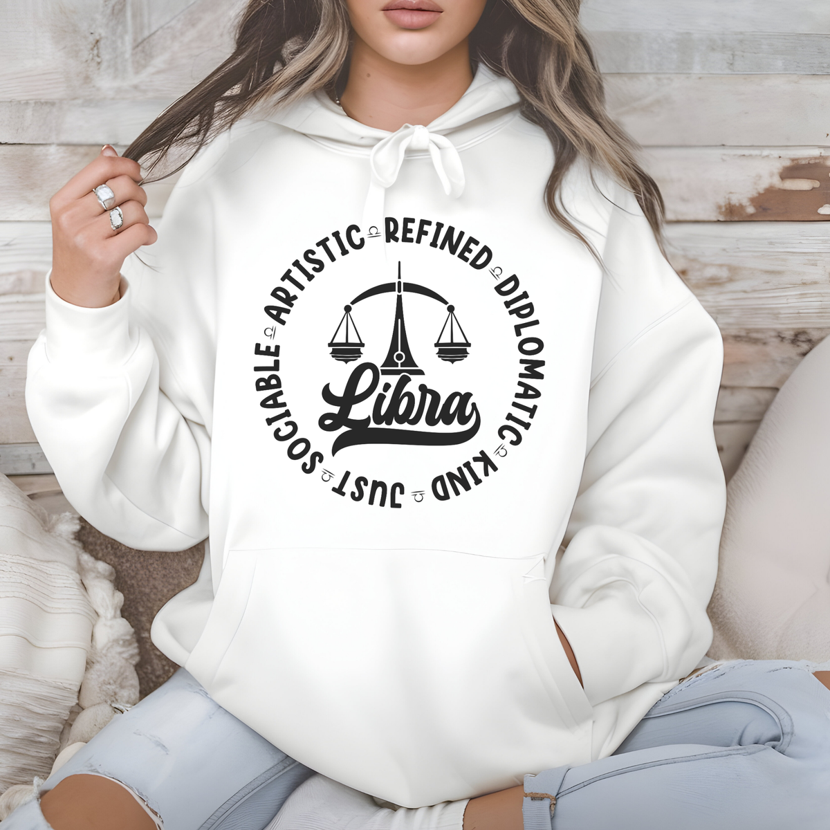 Woman wearing a white crewneck t-shirt featuring a black circular Libra zodiac graphic with a scales illustration, the word "Libra," and trait keywords like artistic, refined, diplomatic, kind, just, and sociable.