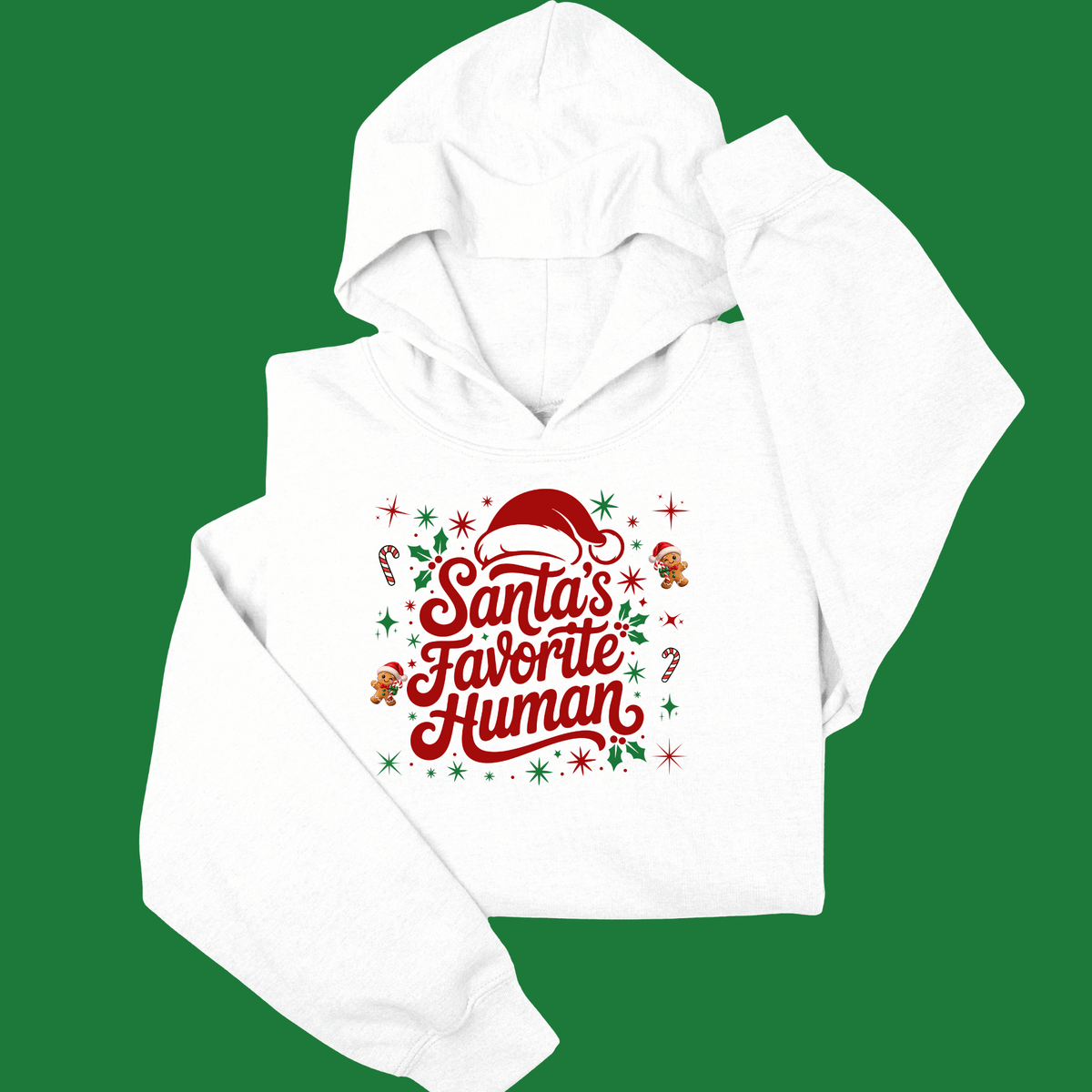 Folded sand crewneck sweatshirt with 'Santa's Favorite Human' red lettering, Santa hat, and gingerbread graphics on a neutral wooden background.