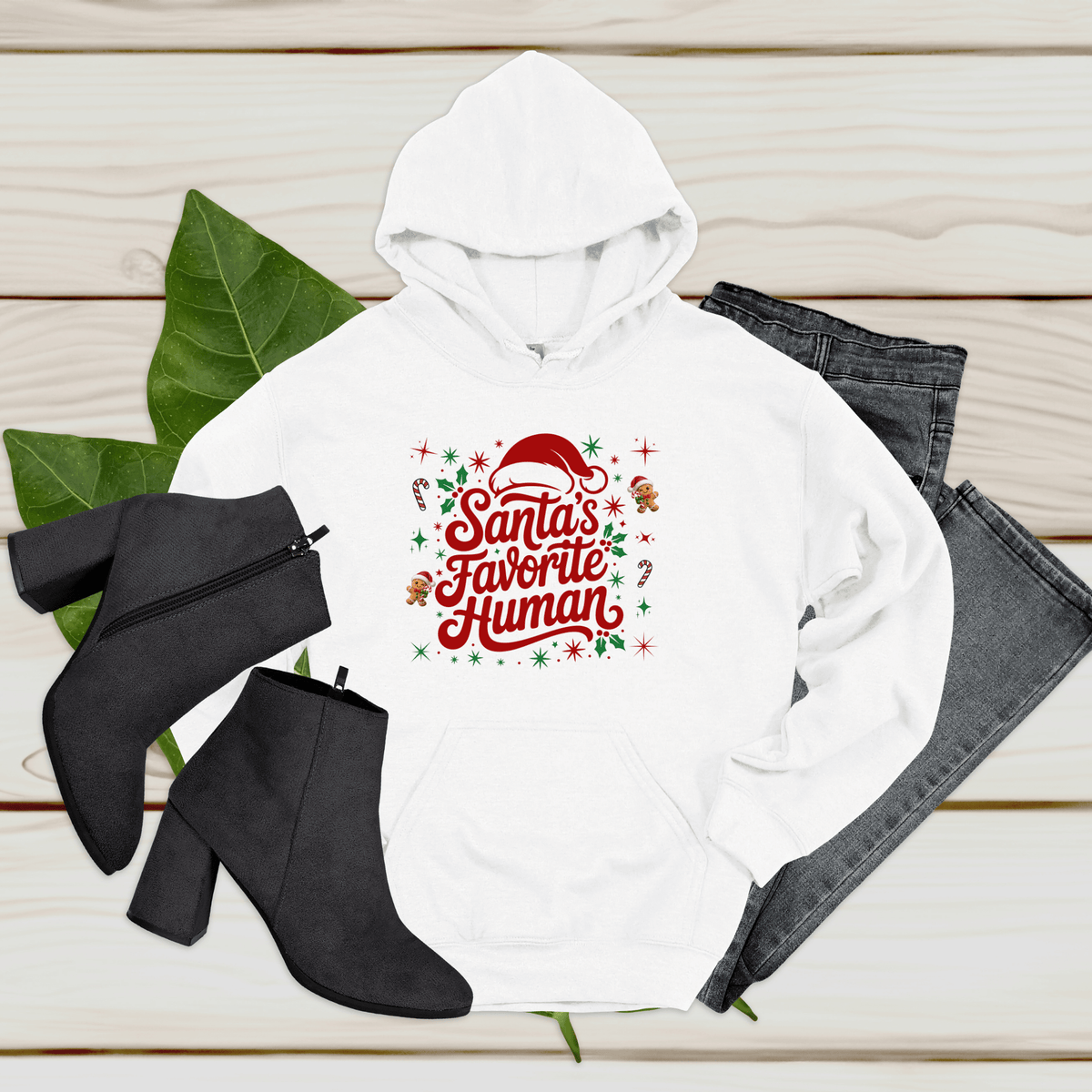 Folded sand crewneck sweatshirt with 'Santa's Favorite Human' red lettering, Santa hat, and gingerbread graphics on a neutral wooden background.