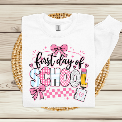 A white long-sleeve t-shirt with a 'first day of SCHOOL' graphic, laid flat on a woven straw mat over a light wood plank background. The graphic features pink bows, patterned letters, and school-themed illustrations.