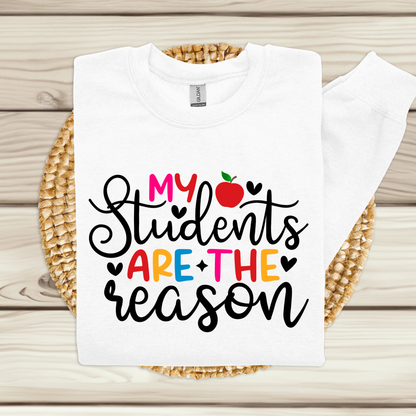 A white long-sleeve sweatshirt laid flat on a woven round mat over a light wood plank surface. The sweatshirt has a colorful graphic print reading “My Students Are The Reason” with a red apple icon and small heart accents.