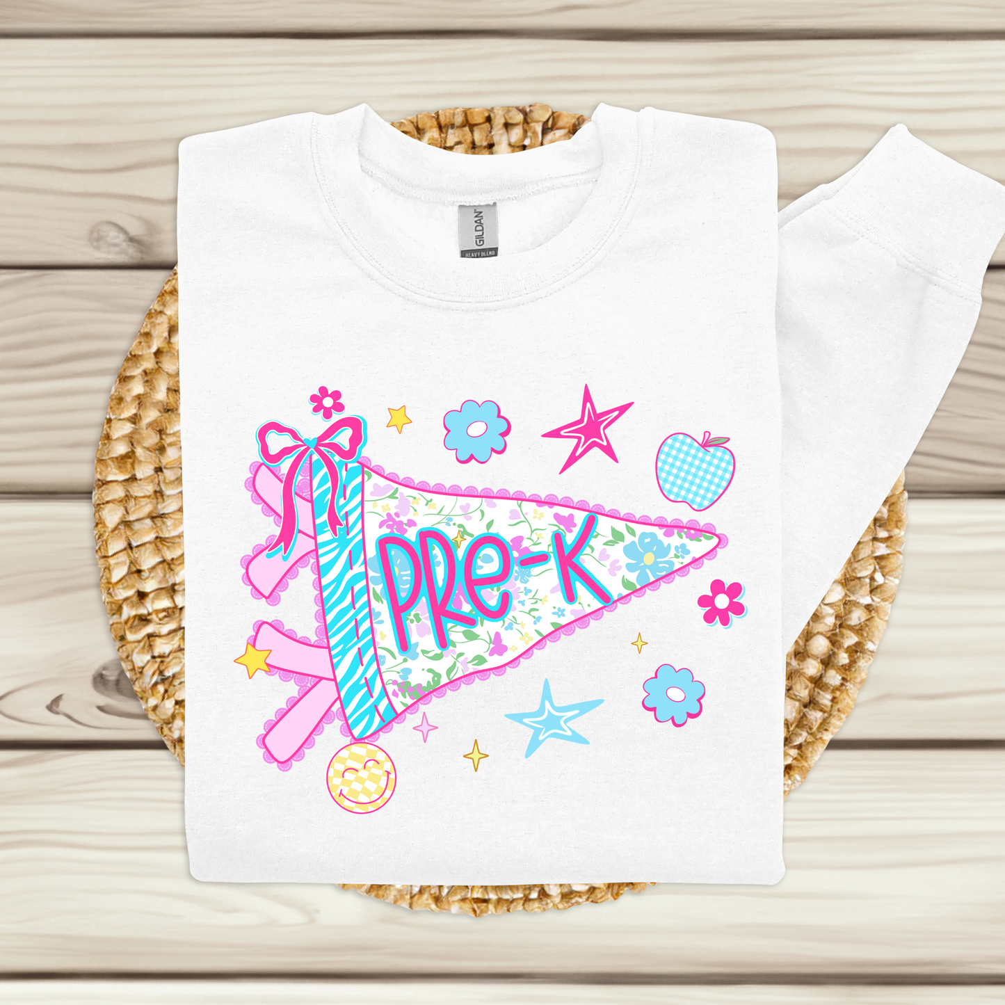 A white long-sleeve sweatshirt laid flat on a woven round mat over a light wood plank surface. The front has a colorful graphic of a pink and blue striped pennant flag with the word 'Pre-K' in purple, surrounded by flowers, stars, and an apple.