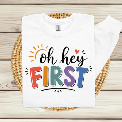 A white crewneck sweatshirt laid flat on a woven round mat over a light wood plank surface. The front graphic reads 'oh hey FIRST' in black script and colorful block letters, with a small red heart and sun illustration.