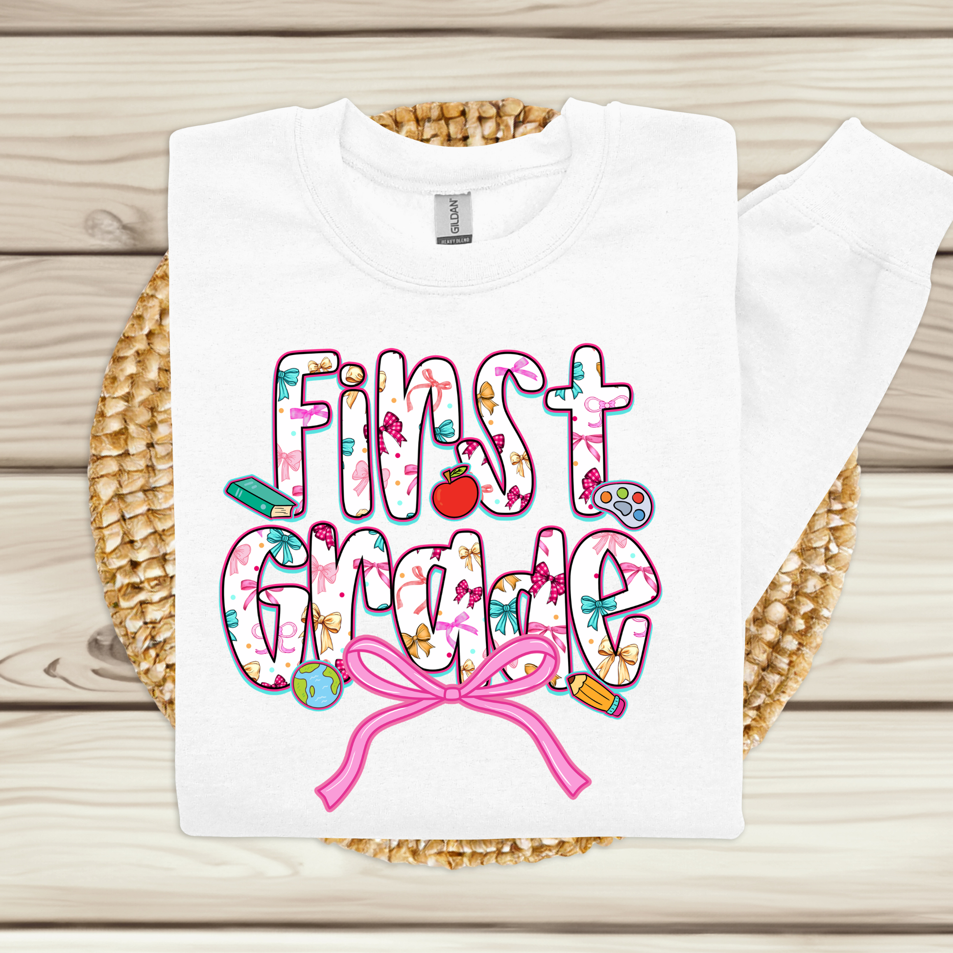 A white long-sleeve sweatshirt laid flat on a woven round mat over a light wood plank background. The front has a colorful 'First Grade' graphic with decorative icons and a pink ribbon motif.