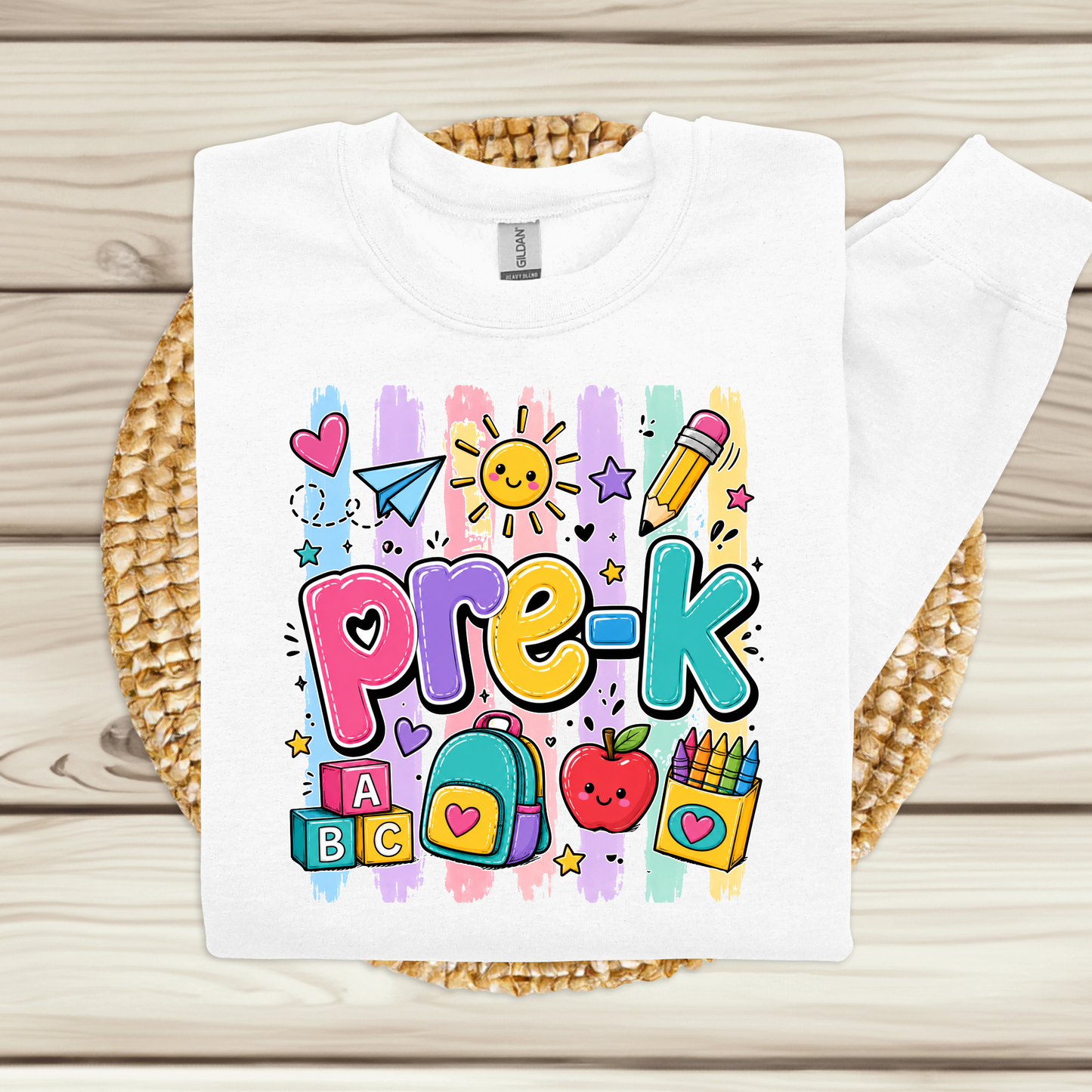 A white long-sleeve sweatshirt laid flat on a woven round mat over a light wood plank surface. The sweatshirt has colorful striped letters spelling 'PRE-K' in yellow, green, blue, orange, and red, with 'Teacher Name' printed below in black script.