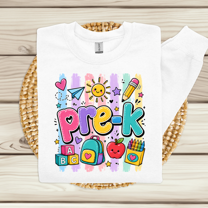 A white long-sleeve sweatshirt laid flat on a woven round mat over a light wood plank surface. The sweatshirt has colorful striped letters spelling 'PRE-K' in yellow, green, blue, orange, and red, with 'Teacher Name' printed below in black script.