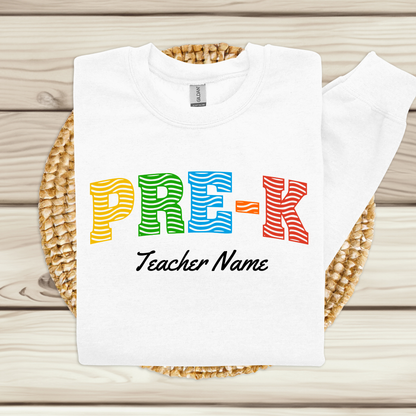 A white long-sleeve sweatshirt laid flat on a woven round mat over a light wood plank surface. The sweatshirt has colorful striped letters spelling 'PRE-K' in yellow, green, blue, orange, and red, with 'Teacher Name' printed below in black script.
