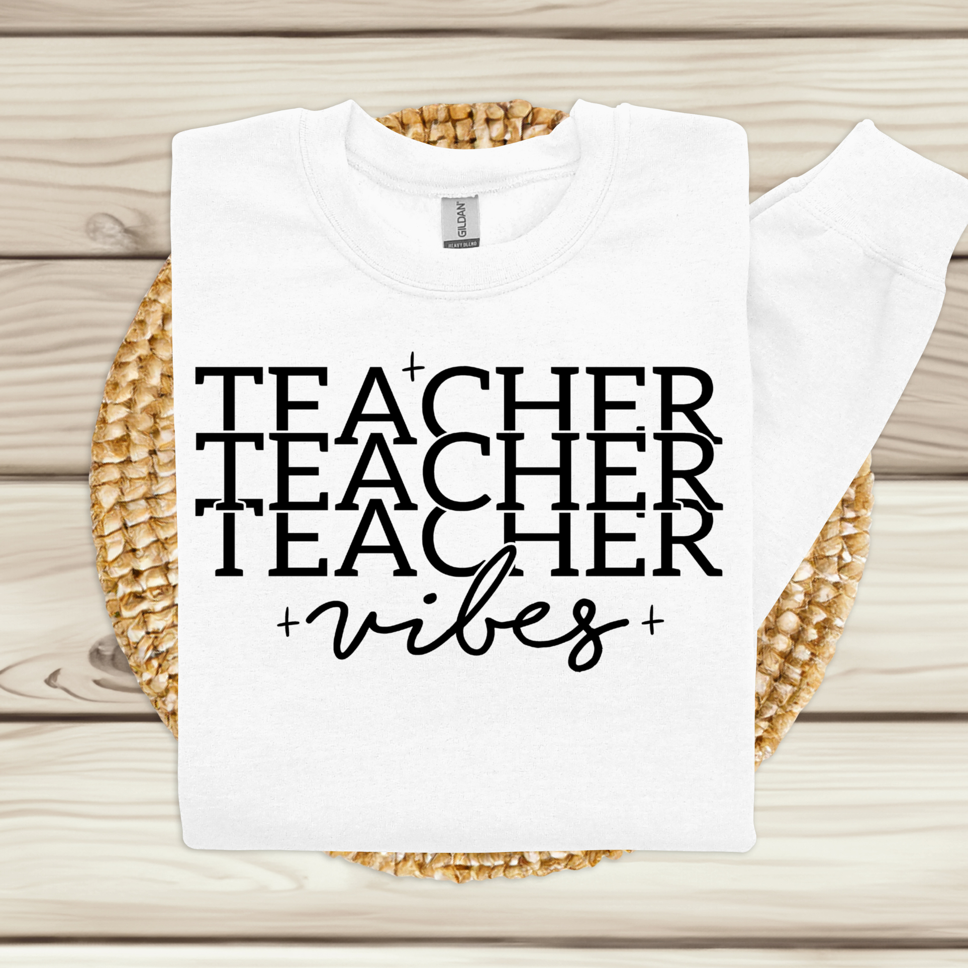 A white crewneck sweatshirt with the text 'TEACHER TEACHER TEACHER +vibes+' printed in black, displayed flat on a woven round mat over a light wood plank background.