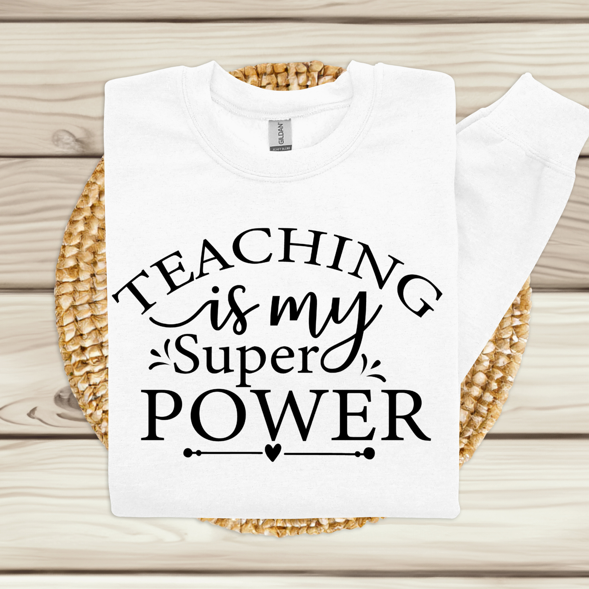 A white crewneck sweatshirt with the text 'TEACHING is my Super POWER' printed in black on the front, displayed flat on a woven round mat over a light wood plank background.