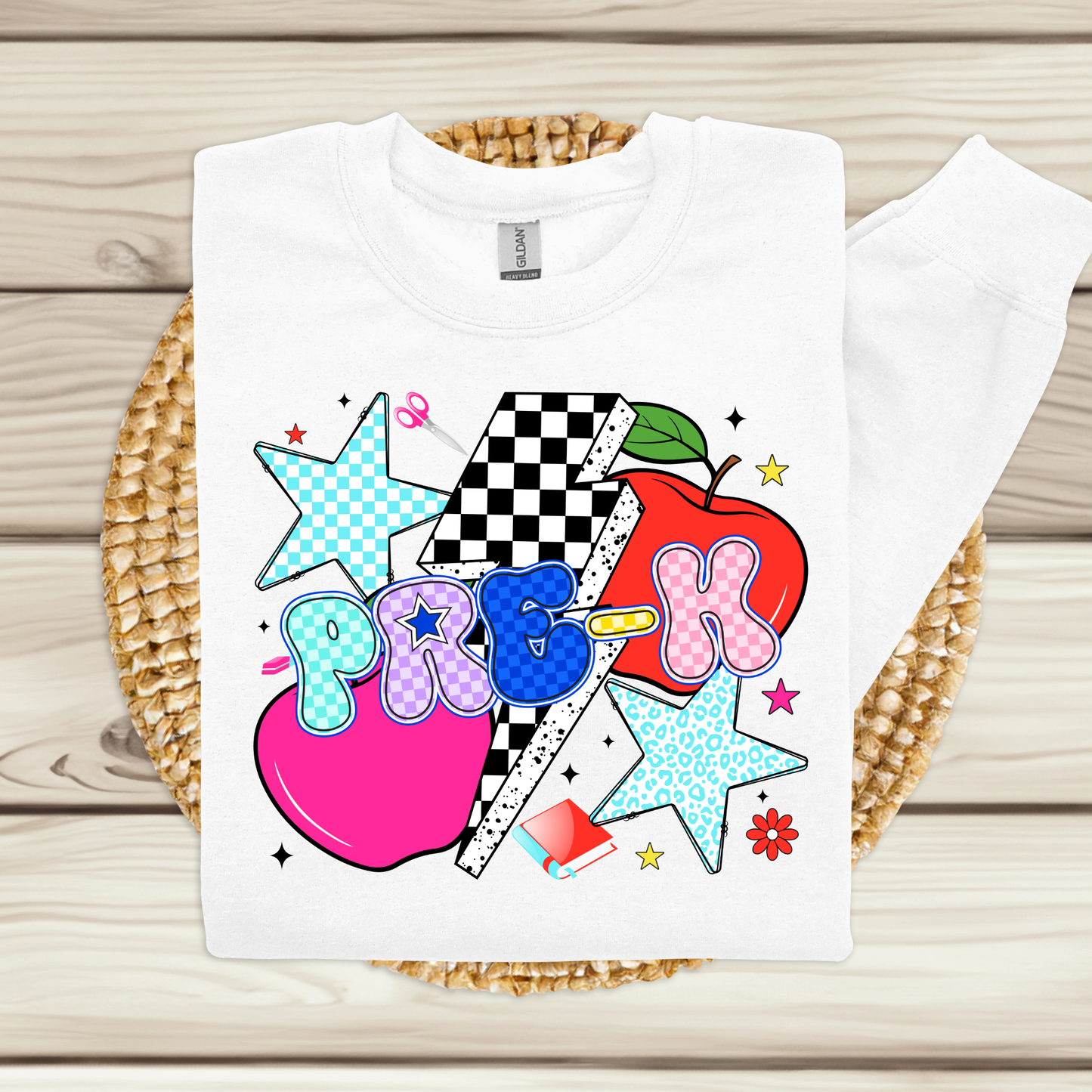 A white long-sleeve sweatshirt laid flat on a woven round mat over a light wood plank background. The front has a colorful school-themed graphic with two red apples, blue patterned stars, a black-and-white checkerboard flag, and the word 'PRIDE' in stylized letters.