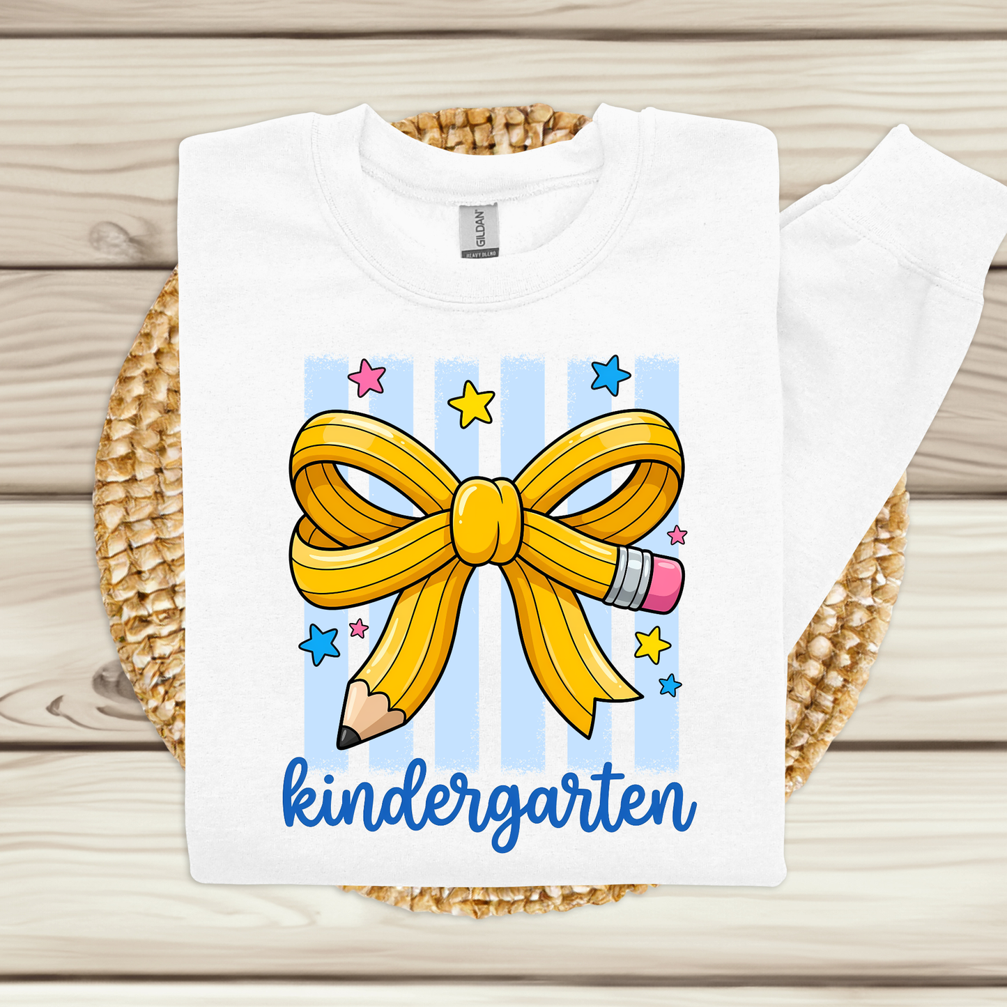 A white long-sleeve t-shirt with a colorful graphic of a yellow pencil bow, blue and pink stars, and the word 'kindergarten' in blue script, displayed on a woven mat over a light wood background.