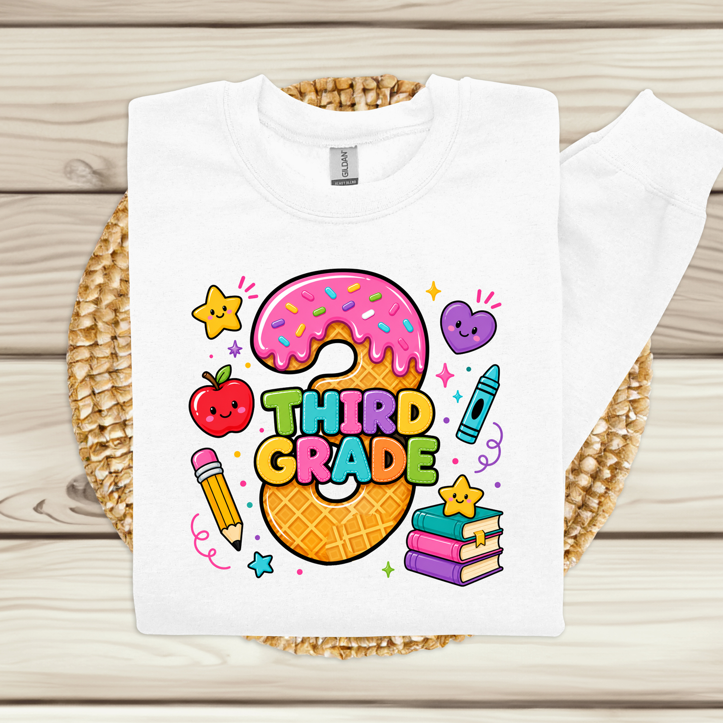 A white long-sleeve sweatshirt laid flat on a woven round mat over a light wood plank surface. The sweatshirt features a colorful school-themed graphic with a donut, the words 'THIRD GRADE,' stars, a heart, a pencil, a crayon, and books.