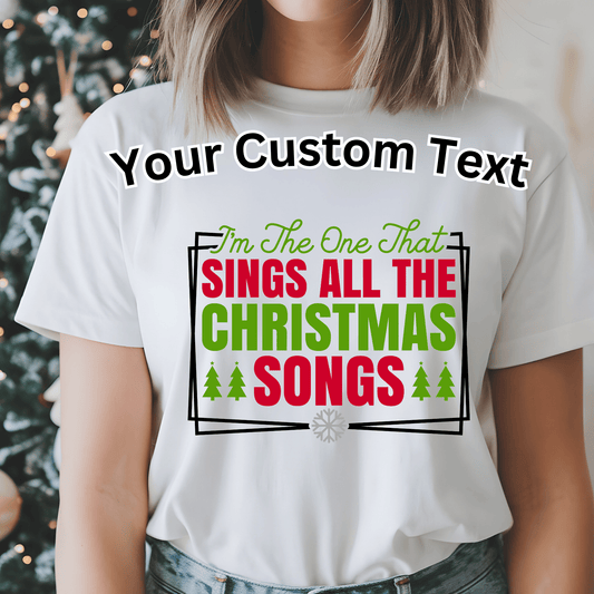 A woman wearing a white short-sleeve graphic T-shirt with the phrase 'Your Custom Text' and a Christmas-themed slogan printed on the front. The shirt is shown on a model in a softly blurred indoor setting with holiday lights and a tree in the background.