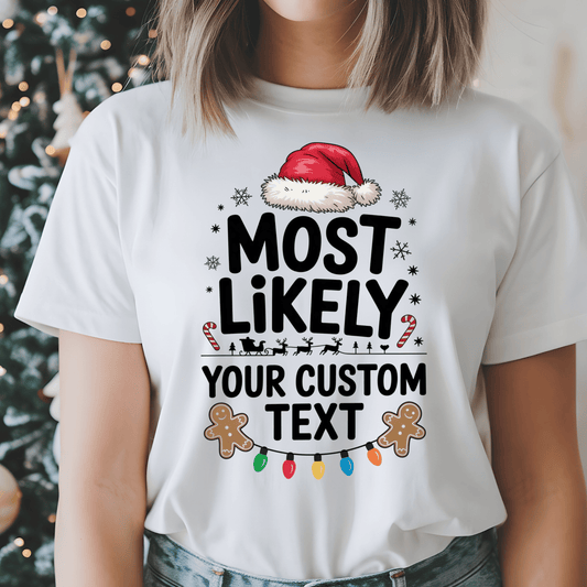 A woman wearing a white graphic t-shirt with a Christmas design that reads 'MOST LIKELY YOUR CUSTOM TEXT' with a Santa hat, gingerbread cookies, candy canes, and lights. The background shows a blurred Christmas tree.