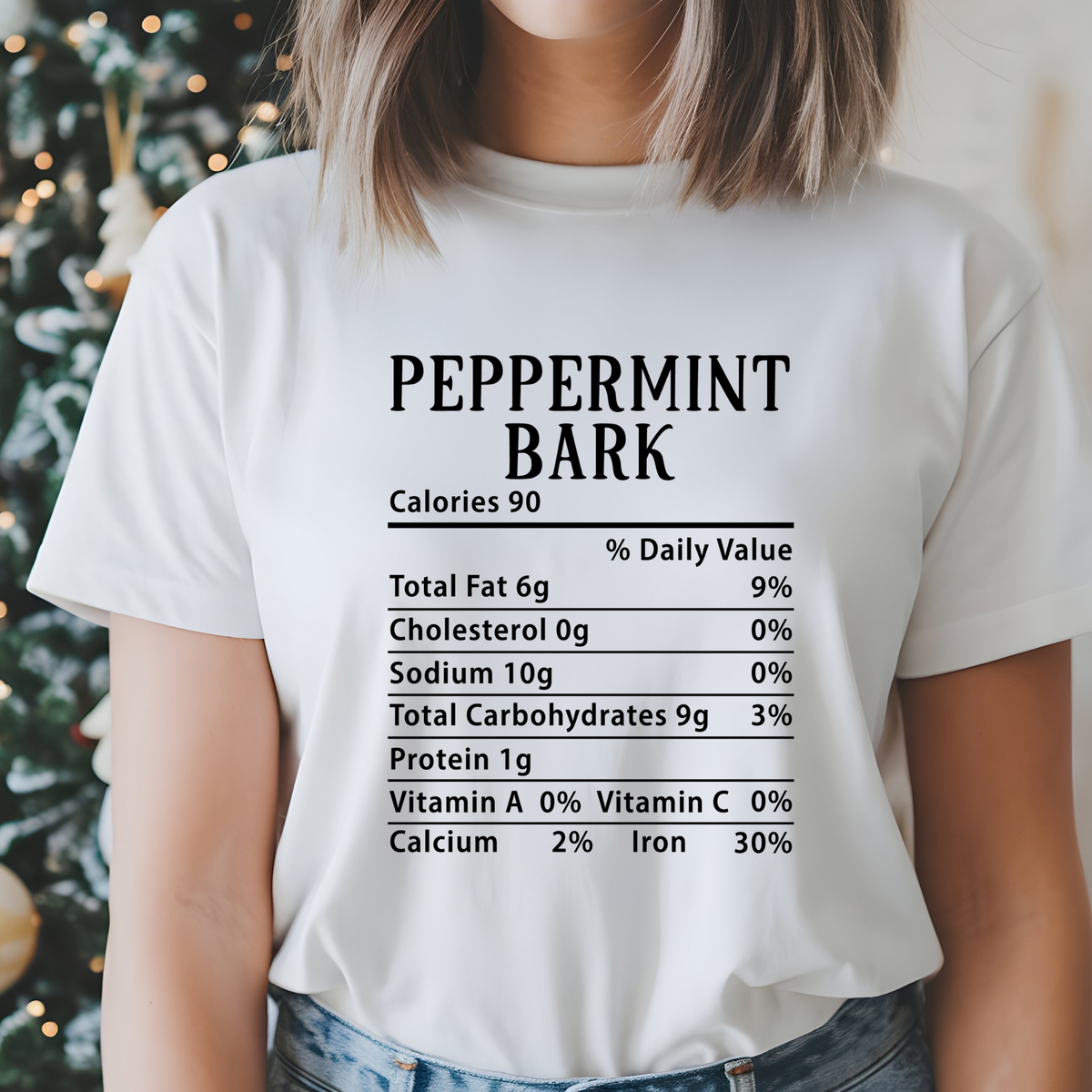 A person wearing a white graphic T-shirt with a black nutrition-style label reading 'PEPPERMINT BARK' and fake nutrient rows, photographed indoors with a blurred holiday tree in the background.