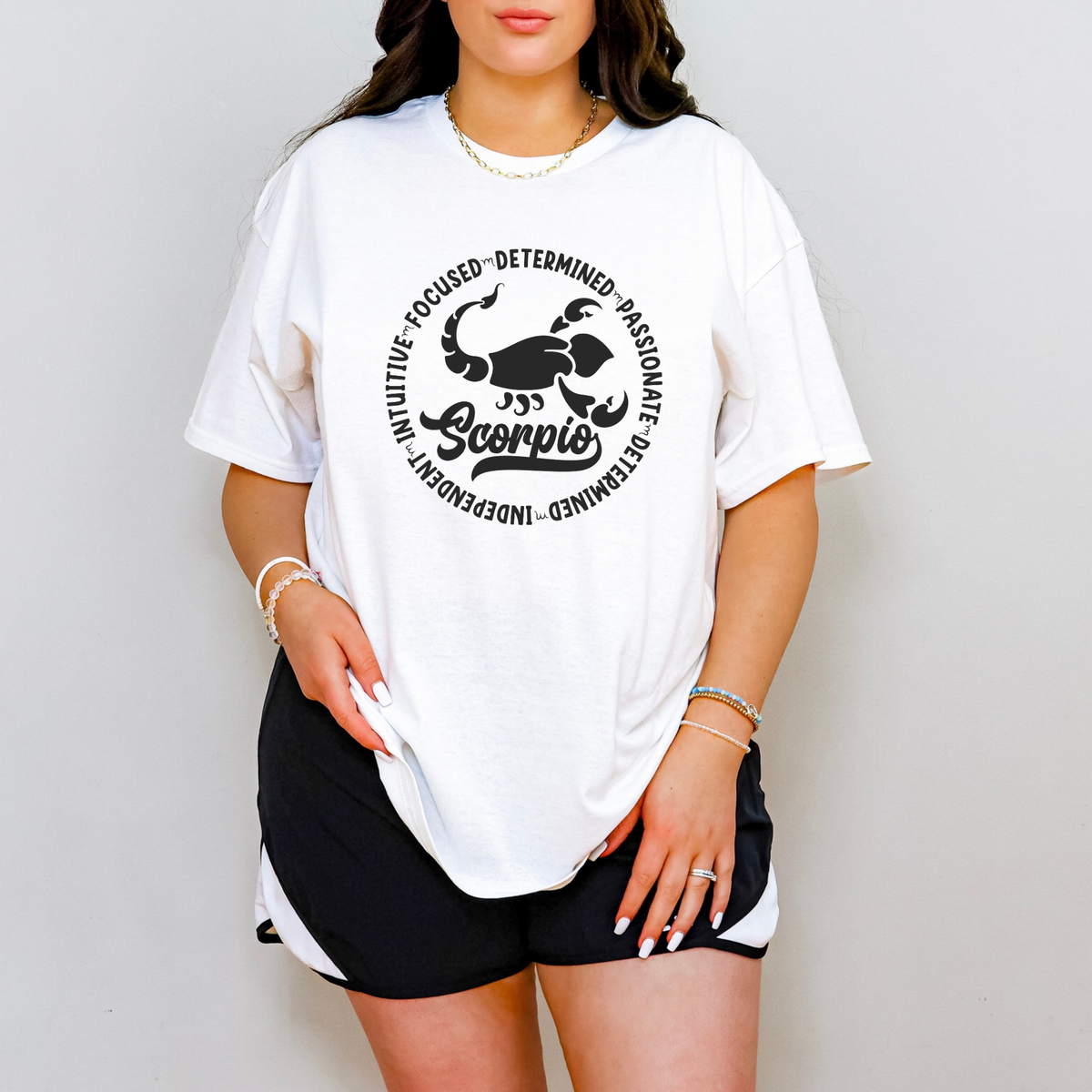 Woman wearing a white tshirt featuring a white circular Scorpio zodiac graphic with a scorpion illustration, the word "Scorpio," and trait keywords like intuitive, focused, determined, passionate, and independent.