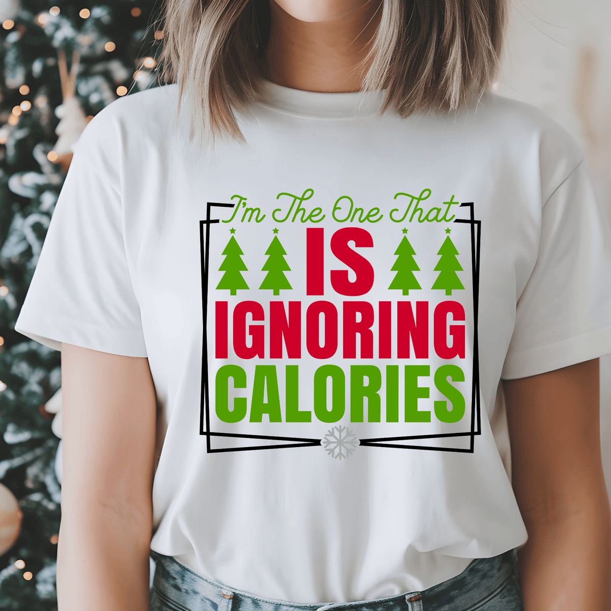 A woman wearing a white graphic t-shirt with the text 'I'm The One That IS IGNORING CALORIES' in green and red, with green Christmas trees, set against a blurred holiday background.