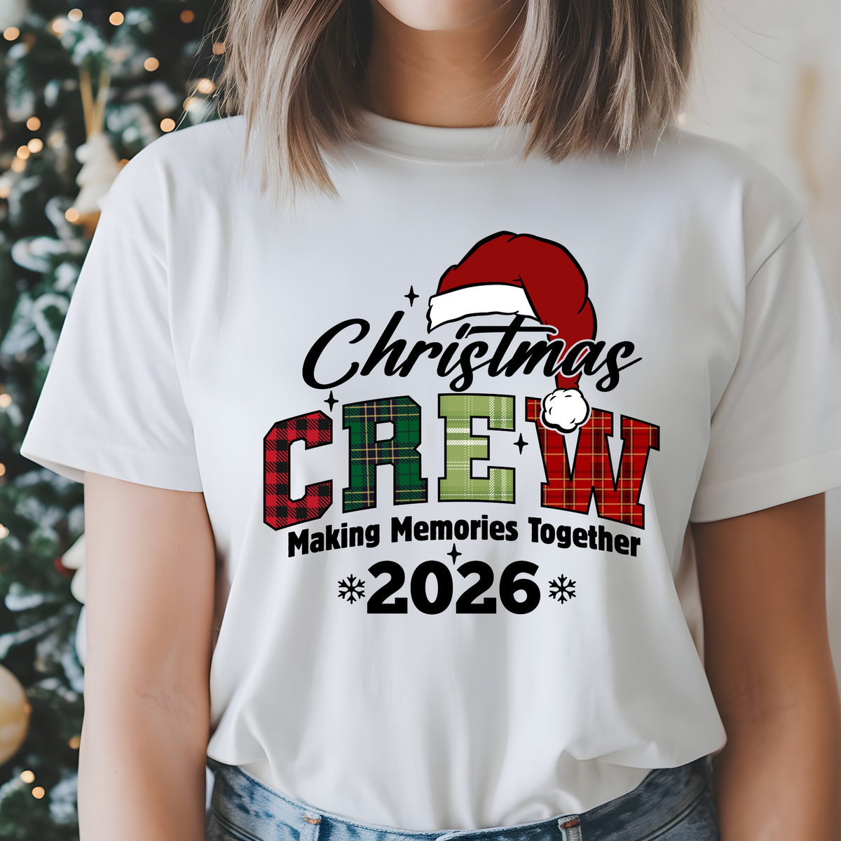 A woman wearing a white graphic t-shirt with a 'Christmas Crew 2026' design featuring a Santa hat and plaid lettering, set against a blurred Christmas tree background.