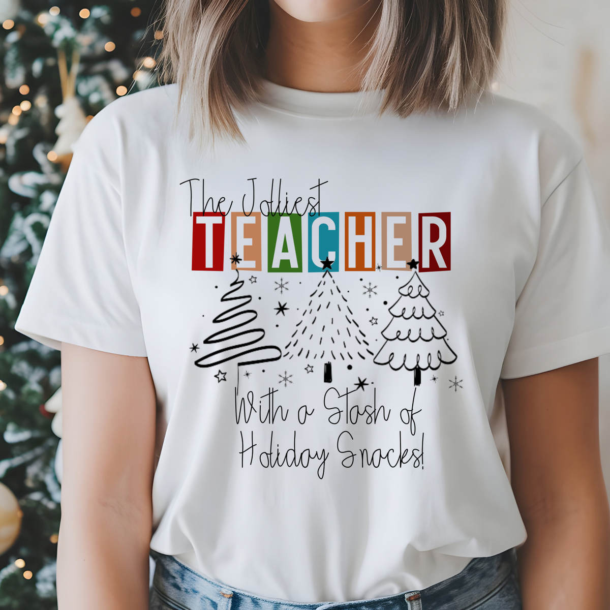 A woman wearing a white graphic t-shirt with a Christmas-themed design that reads 'The Jollyest TEACHER With a Stash of Holiday Snacks!' in colorful block letters and black line art of Christmas trees, set against a softly blurred holiday background.