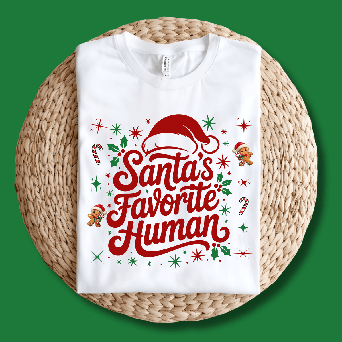 Folded sand crewneck sweatshirt with 'Santa's Favorite Human' red lettering, Santa hat, and gingerbread graphics on a neutral wooden background.