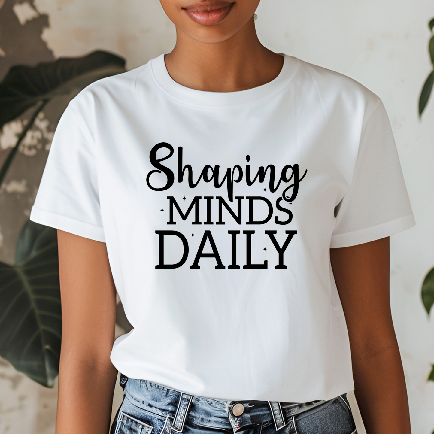 A woman wearing a white short-sleeve graphic T-shirt with the phrase 'Shaping MINDS DAILY' printed in black across the chest, paired with blue jeans in a softly lit indoor setting with blurred plants in the background.