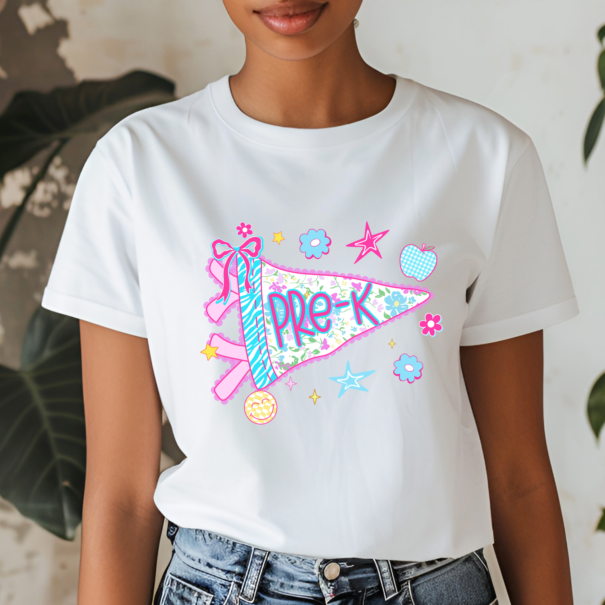 A woman wearing a white short-sleeve t-shirt with a colorful 'Pre-K' pennant graphic, paired with blue jeans. The background is a clean indoor setting with a white wall and some green plants.