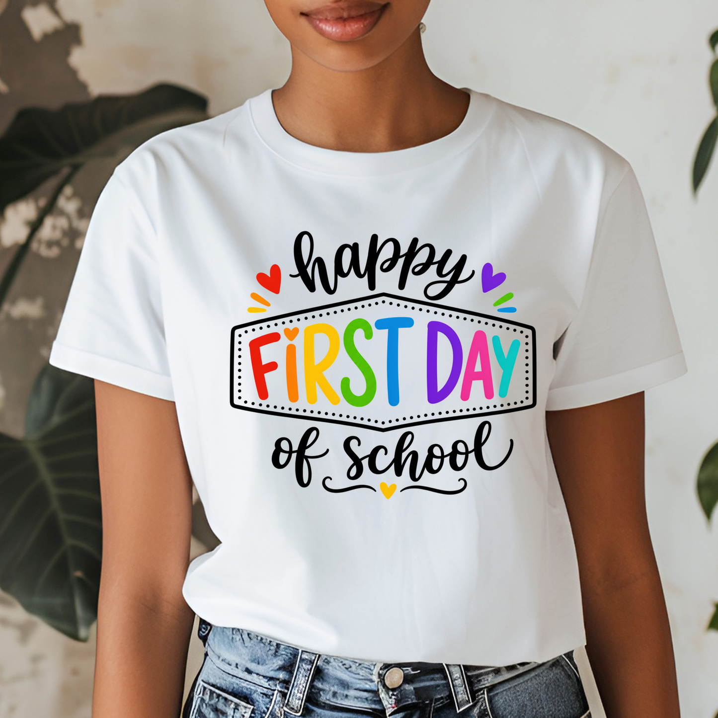 A woman wearing a white graphic t-shirt with a colorful 'First Day of School' print, paired with blue jeans, set against a softly blurred indoor background with plants.