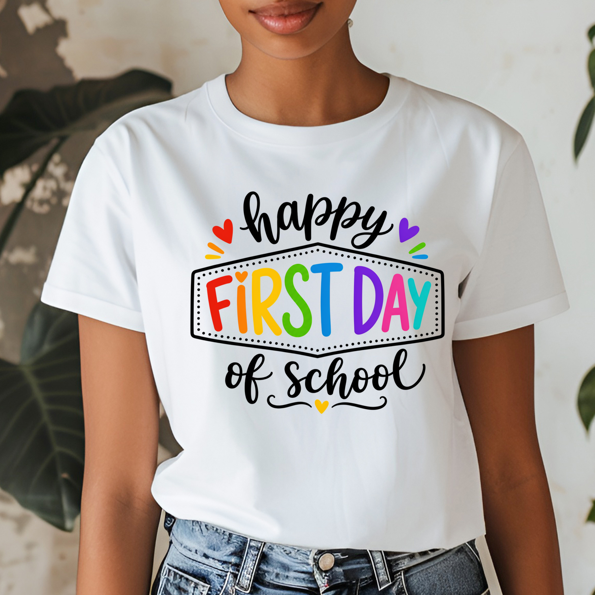A woman wearing a white graphic t-shirt with a colorful 'First Day of School' print, paired with blue jeans, set against a softly blurred indoor background with plants.