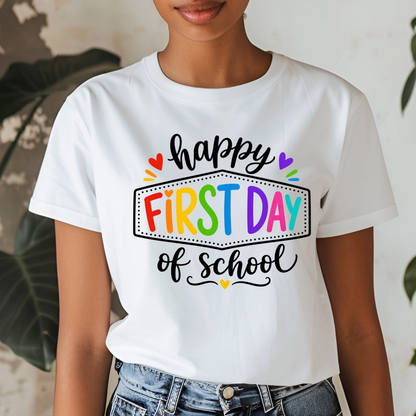 A woman wearing a white graphic t-shirt with a colorful 'First Day of School' print, paired with blue jeans, set against a softly blurred indoor background with plants.