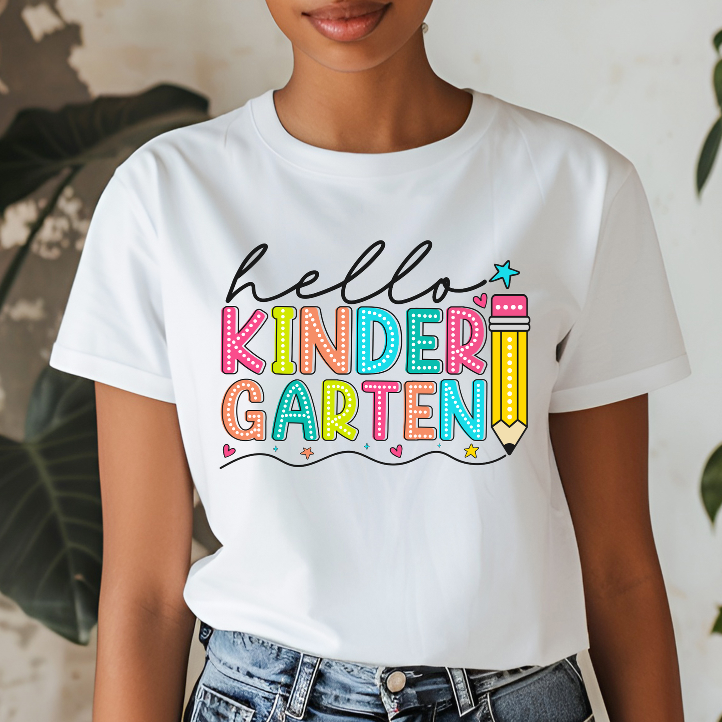 A woman wearing a white short-sleeve graphic T-shirt with the phrase 'hello KINDER GARTEN' in colorful playful lettering, paired with blue jeans. The background is a softly blurred indoor setting with green leaves.