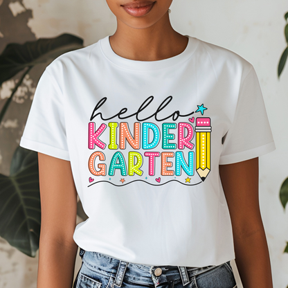 A woman wearing a white short-sleeve graphic T-shirt with the phrase 'hello KINDER GARTEN' in colorful playful lettering, paired with blue jeans. The background is a softly blurred indoor setting with green leaves.