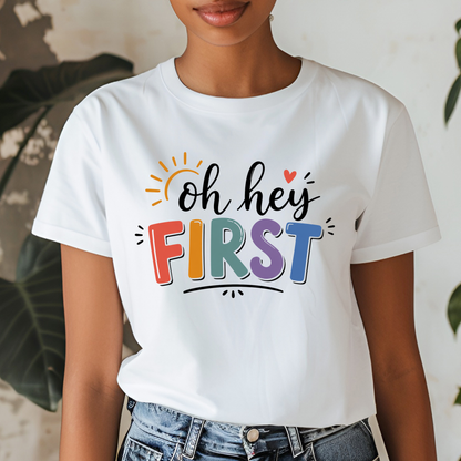 A woman wearing a white short-sleeve graphic T-shirt with the phrase 'hello KINDER GARTEN' in colorful playful lettering, paired with blue jeans. The background is a softly blurred indoor setting with green leaves.