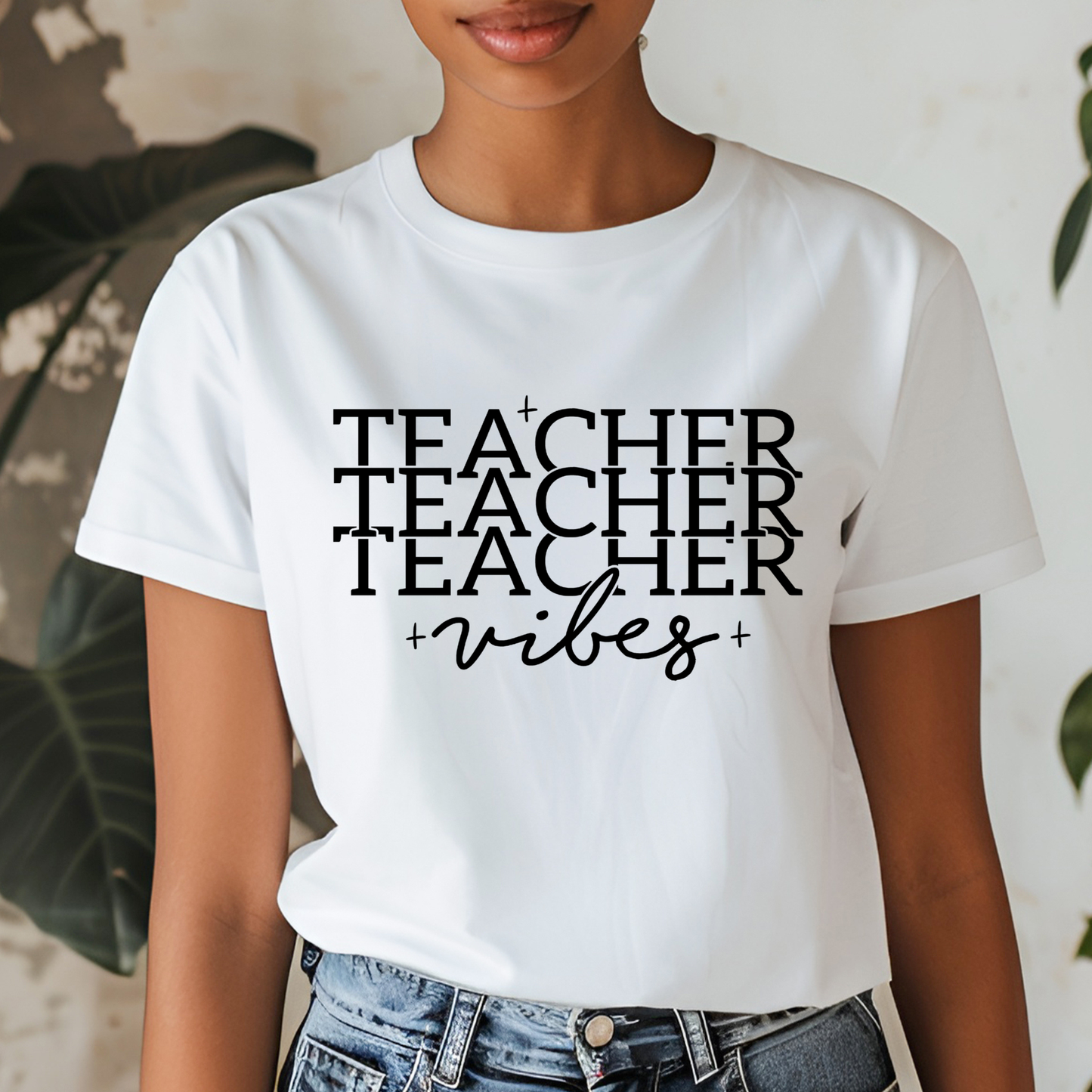 A woman wearing a white short-sleeve graphic T-shirt with the text 'TEACHER TEACHER TEACHER vibes' printed in black across the front, paired with blue jeans.
