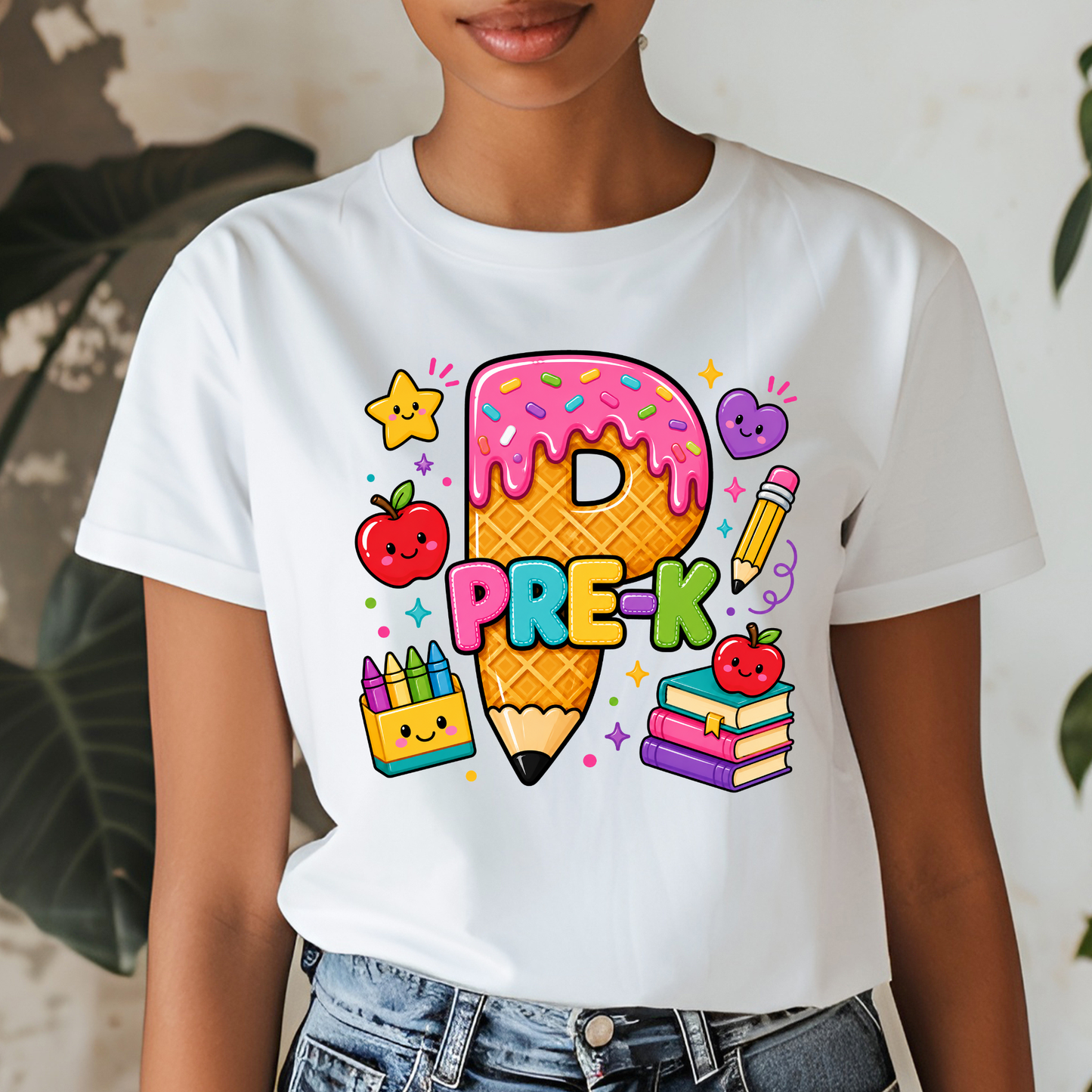 A person wearing a white short-sleeve T-shirt with a colorful cartoon graphic of a pencil-shaped ice cream cone, the word "PRE-K," apples, crayons, and books. The photo is cropped from the lower face to the hips, with jeans visible and a light indoor background with blurred plants.