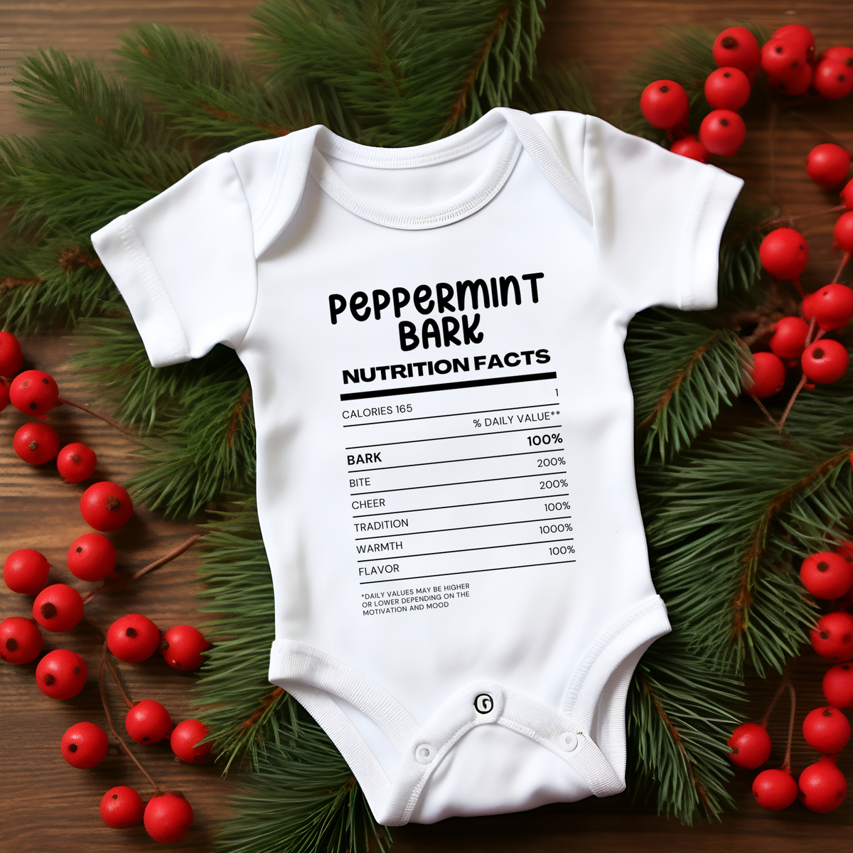 A white short-sleeve baby bodysuit laid flat on a wooden surface, surrounded by green pine branches and red berries. The bodysuit has a black printed nutrition facts label graphic with the text 'PEPPERMINT BARK' and humorous nutrition details.