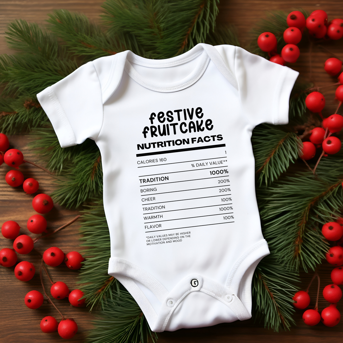 A white short-sleeve baby bodysuit laid flat on a wooden surface, surrounded by green pine branches and red berries. The bodysuit has a black printed graphic styled like a nutrition facts label with the text 'festive fruitcake' and a fake nutrition table.