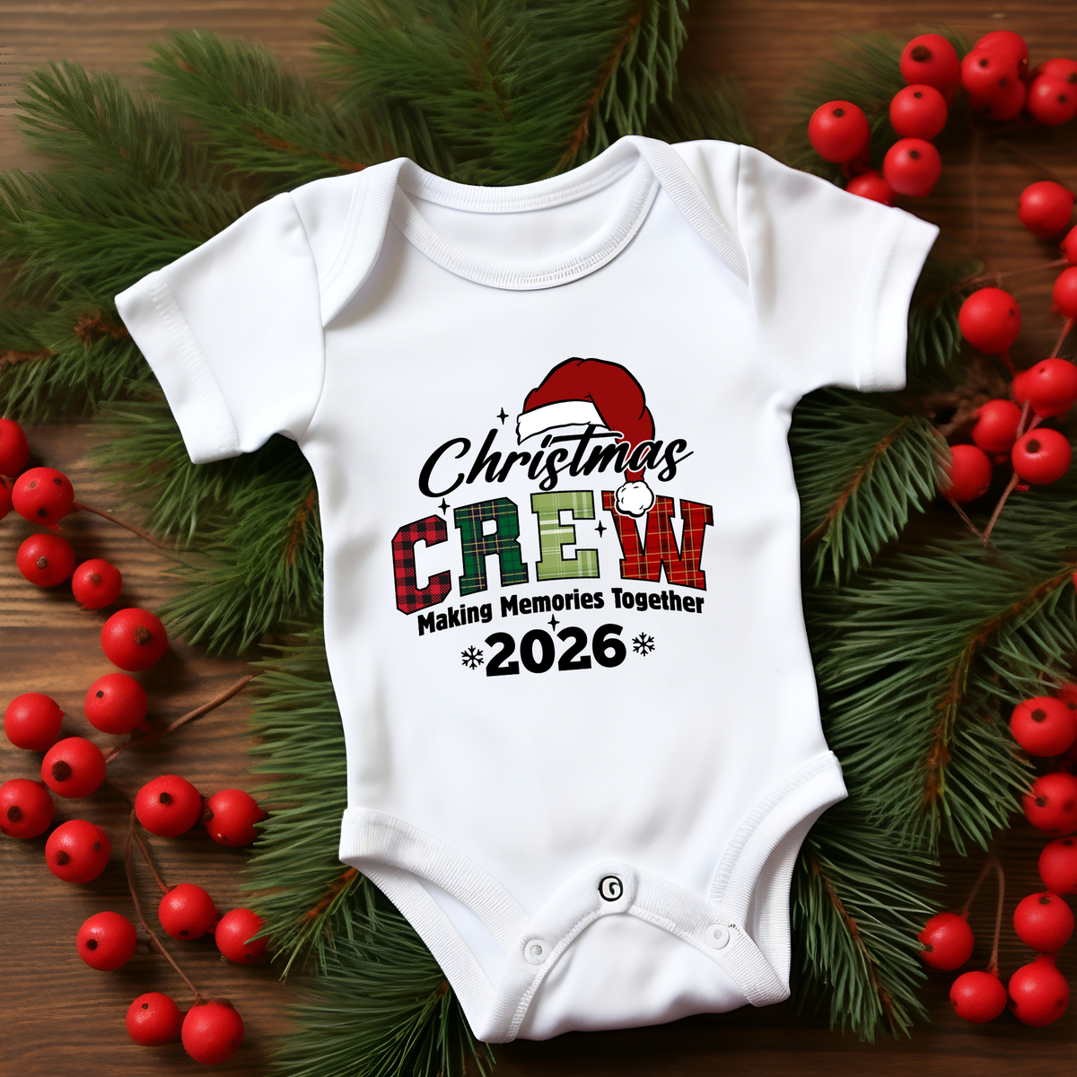A white short-sleeve baby bodysuit with a Christmas-themed graphic reading 'Christmas Crew Making Memories Together 2026,' styled flat on a wooden surface with green pine branches and red berries around it.