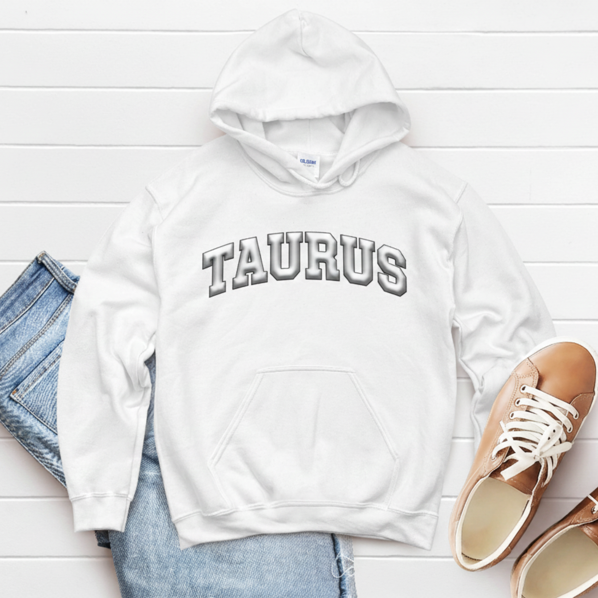 White hoodie with 'TAURUS' text on a white wooden surface with jeans and shoes.