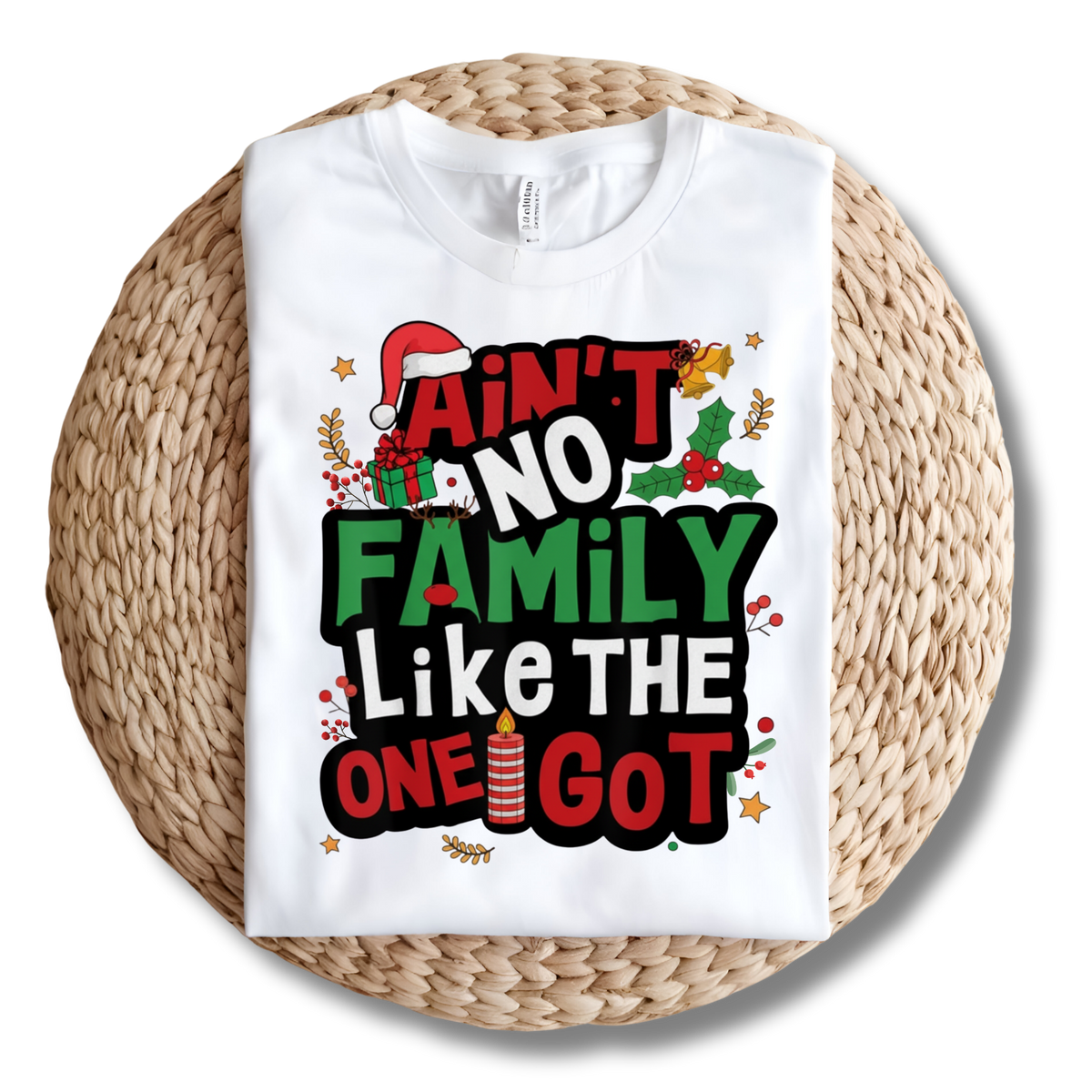 A white short-sleeve T-shirt laid flat on a round woven straw mat, featuring a large Christmas-themed graphic with the phrase “Ain’t No Family Like the One I Got” in red, green, black, and white lettering with holiday illustrations.