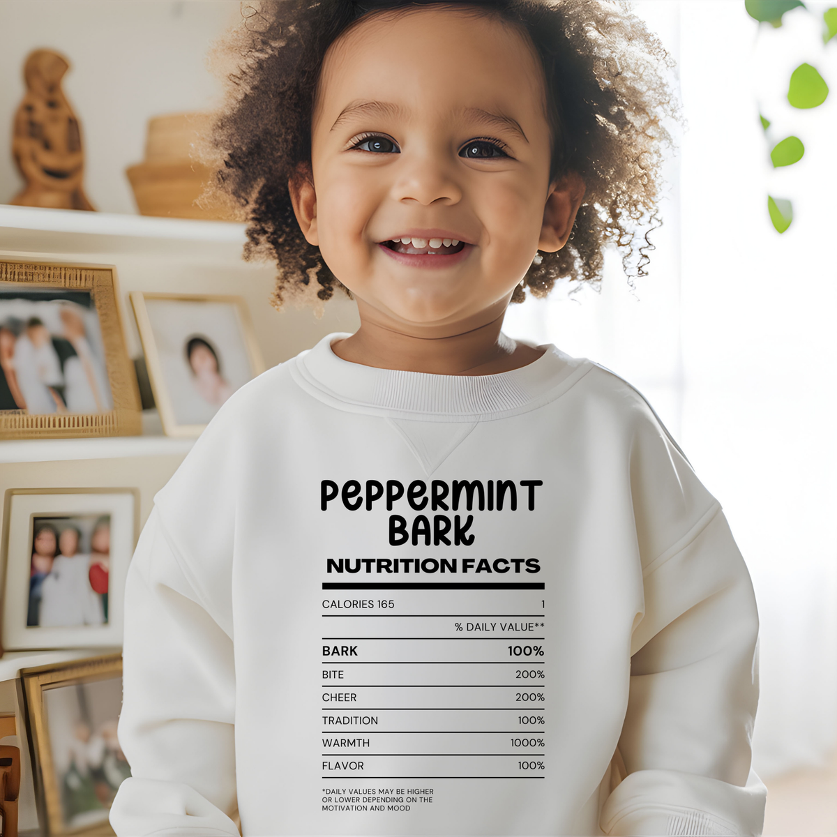 A smiling toddler with curly hair wearing a white sweatshirt with a black 'Peppermint Bark Nutrition Facts' graphic, standing in a bright indoor room with framed photos on a shelf behind them.