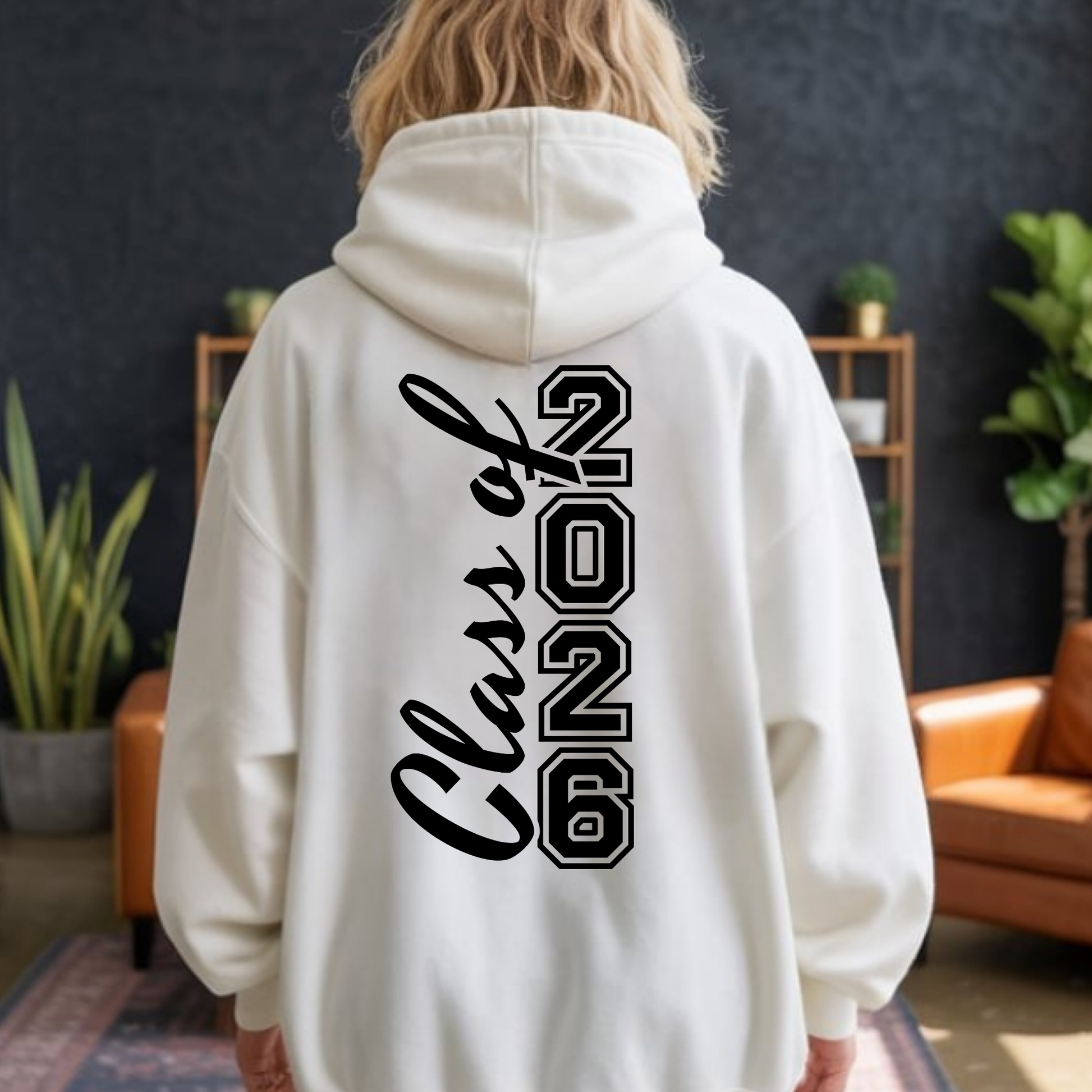 Person wearing a white hoodie with 'Class of 2026' text on the back in a room with plants and furniture.  Senior 2026 Shirt -  Celebrate the Graduating Class of 2026 In Bold Style: Front & Back