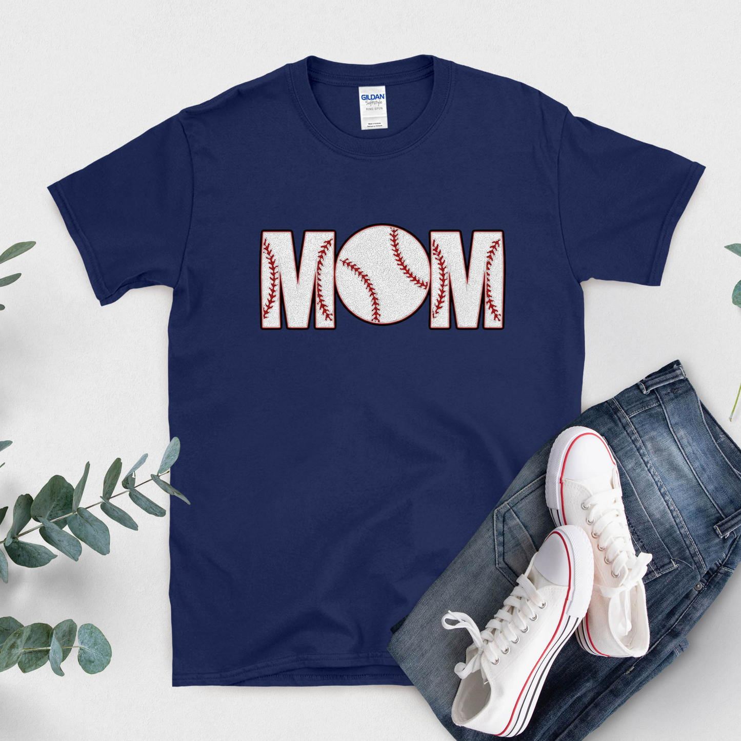 Navy blue t-shirt with 'BASEBALL MOM' design, white sneakers, and jeans on a light gray background.