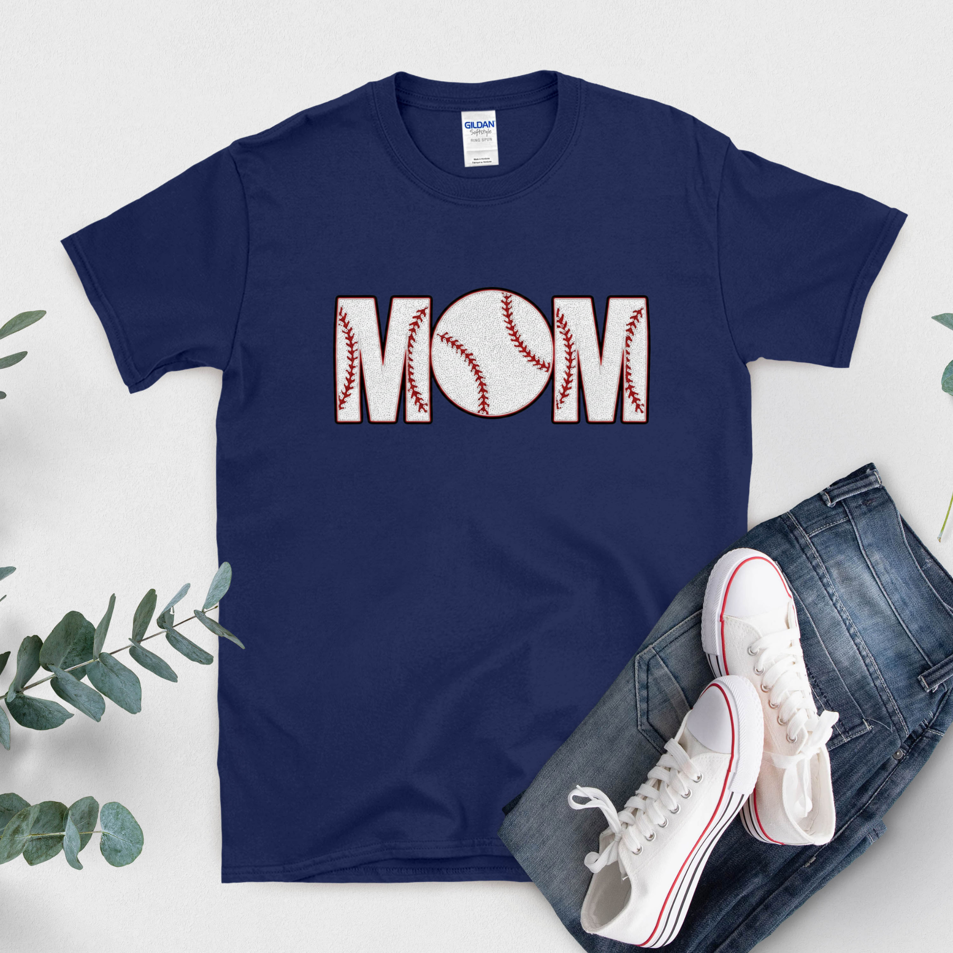 Navy blue t-shirt with 'BASEBALL MOM' design, white sneakers, and jeans on a light gray background.