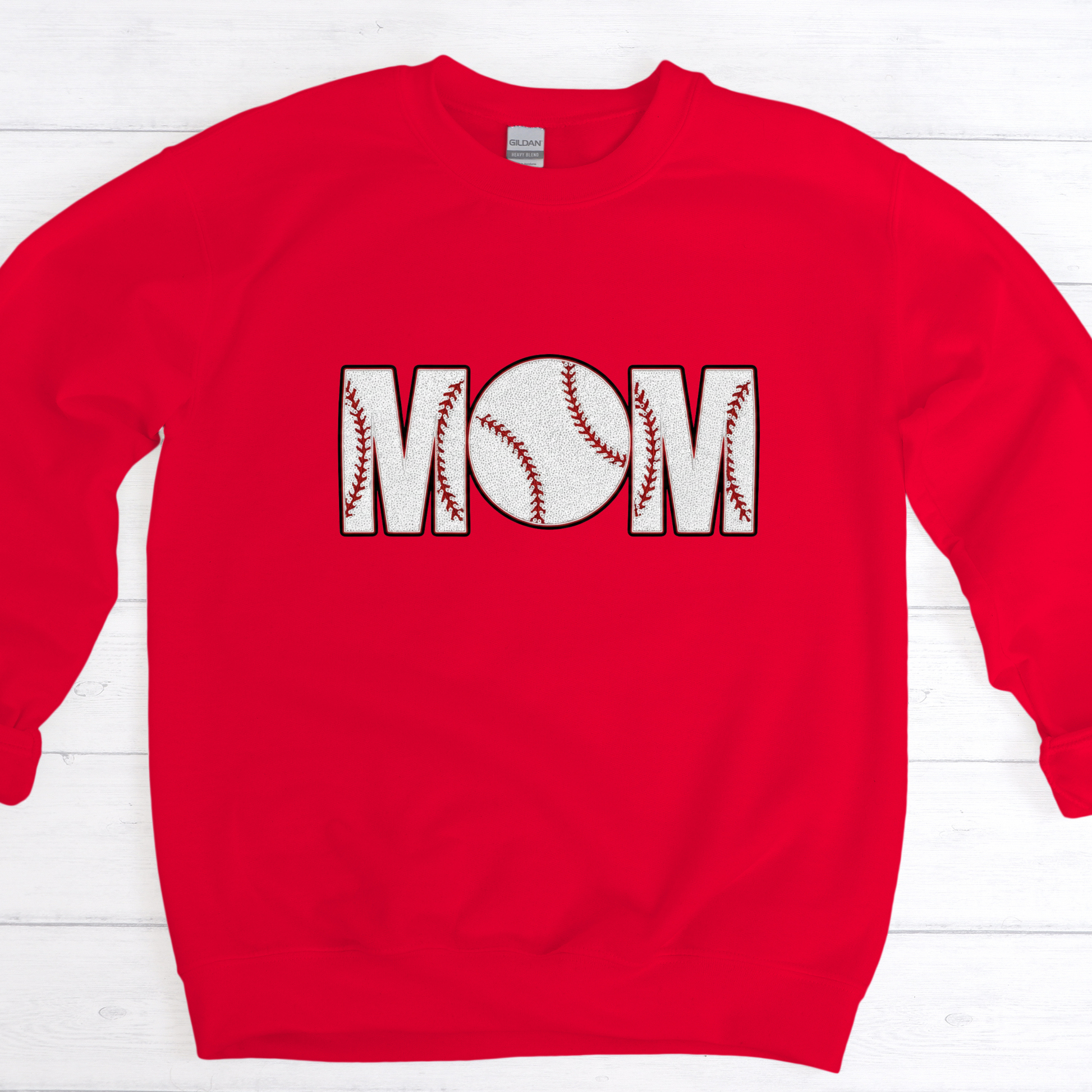 Red sweatshirt with 'Mom' printed on it featuring baseball designs, on a white wooden background.