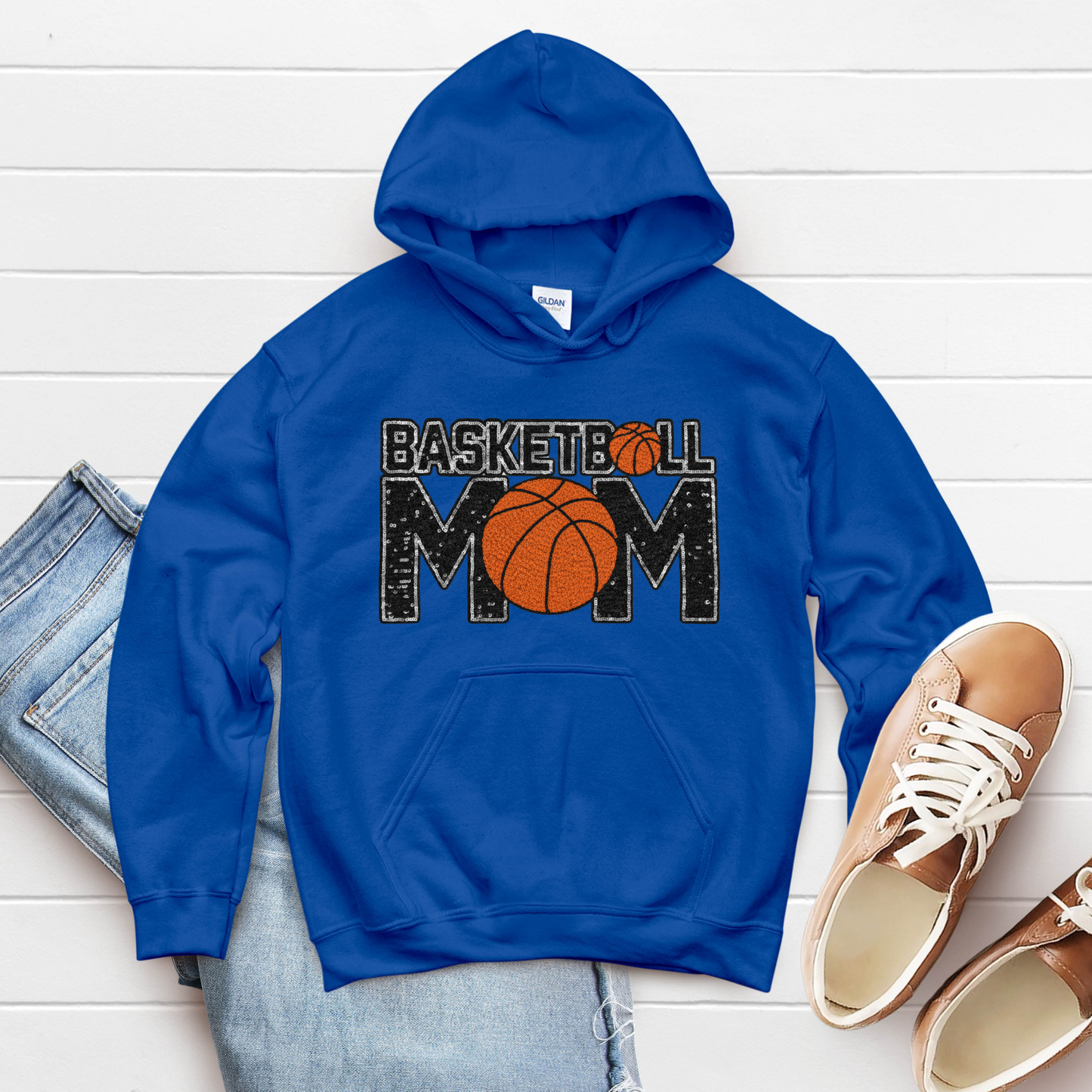 Blue hoodie with 'Basketball Mom' text and basketball graphic on a white wooden surface with jeans and shoes.