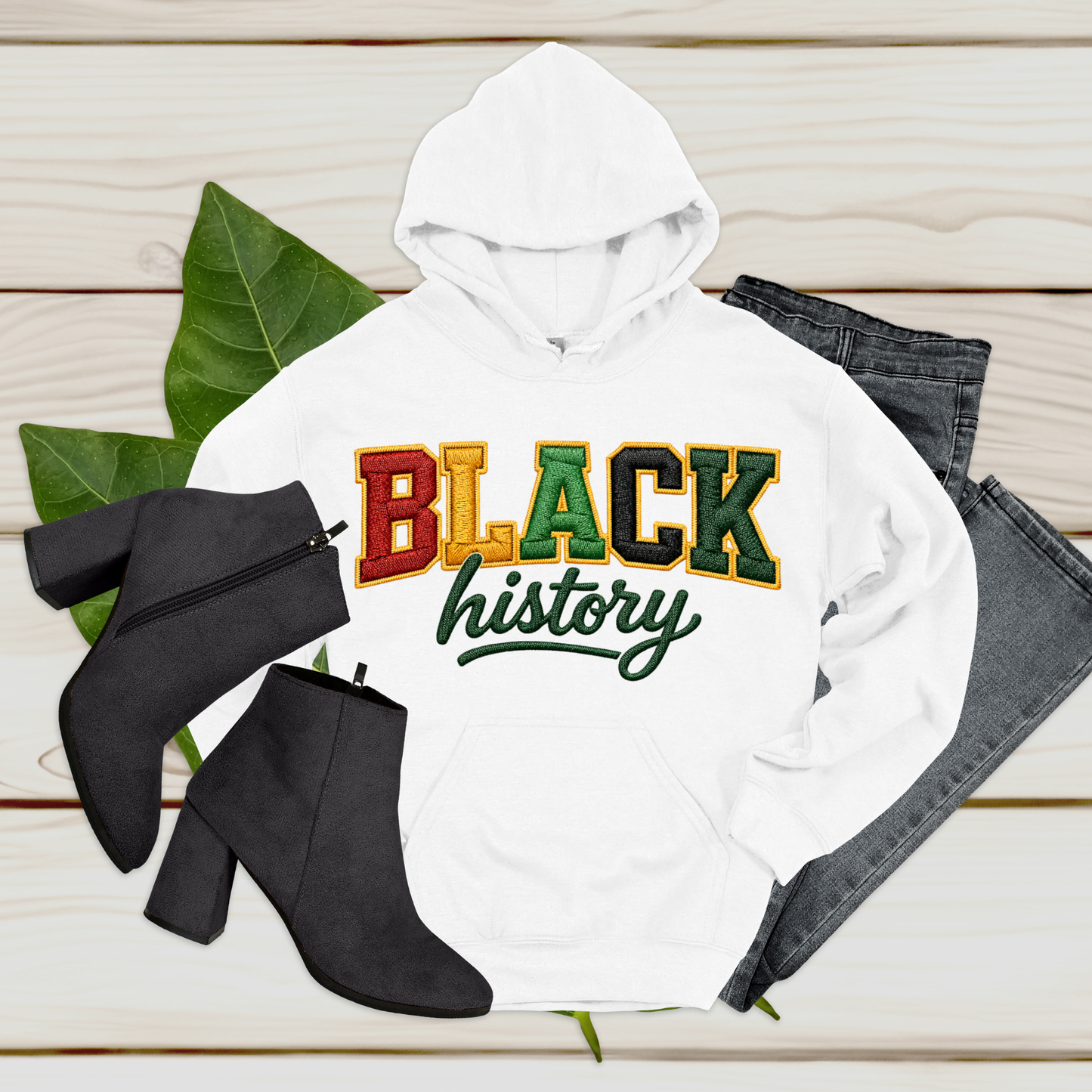 White hoodie with 'BLACK history' text, black ankle boots, and jeans on a wooden surface.