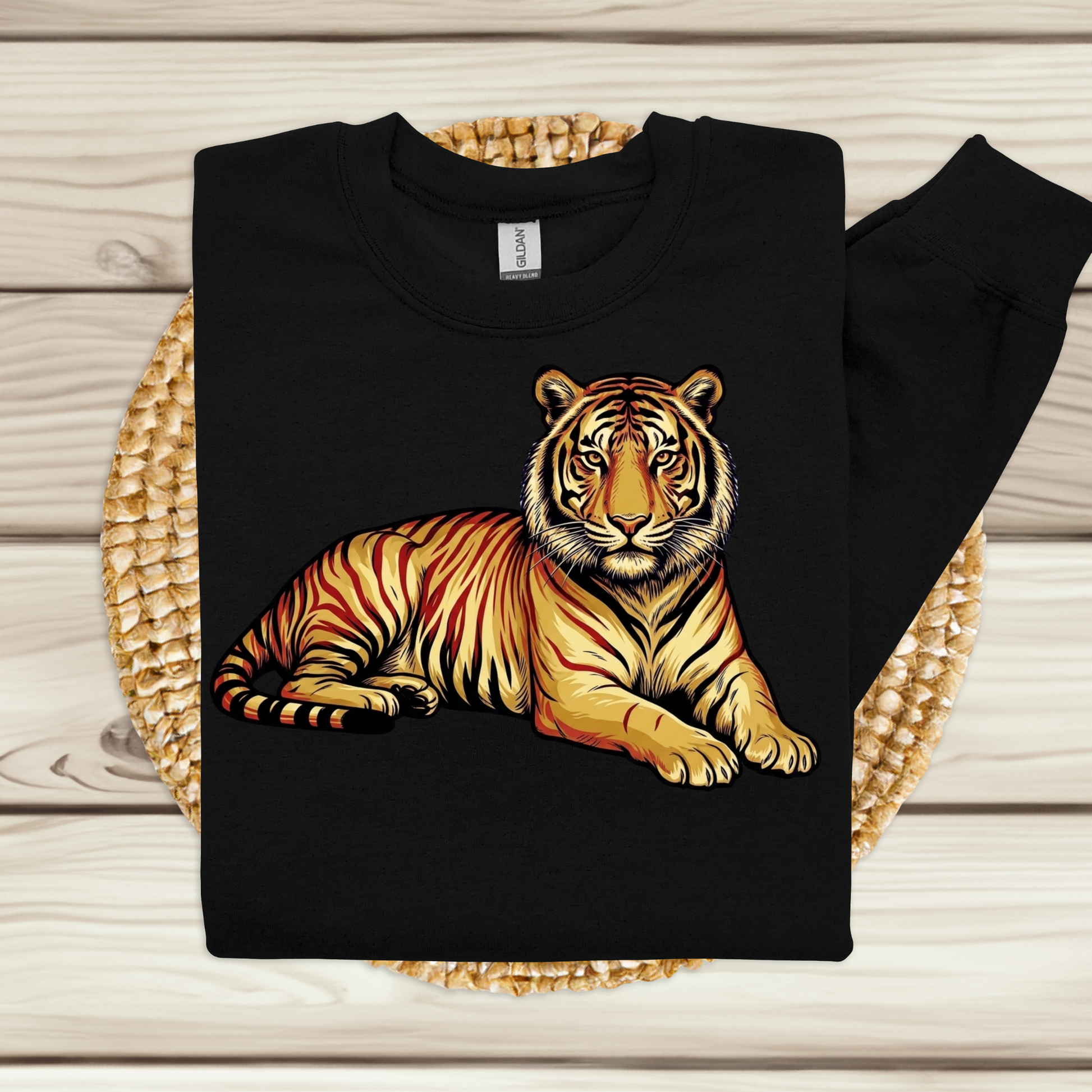 A black long-sleeve crewneck sweatshirt featuring a detailed graphic of a resting tiger in orange, yellow, and black, displayed flat on a woven straw mat over a light wood plank background.